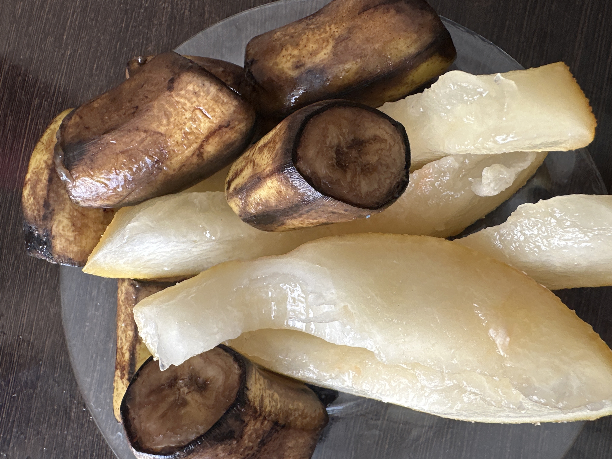 The image shows a close-up, overhead view of a clear glass plate containing several peeled, cooked bananas and pieces of cooked pear. The bananas are browned and appear soft, with some black spots on their skins. The pear pieces are translucent and pale yellow, with a gelatinous texture. The plate is on a dark wooden surface, likely a table. The scene is indoors. There are no people present in the image. The location is not discernible from the image itself.