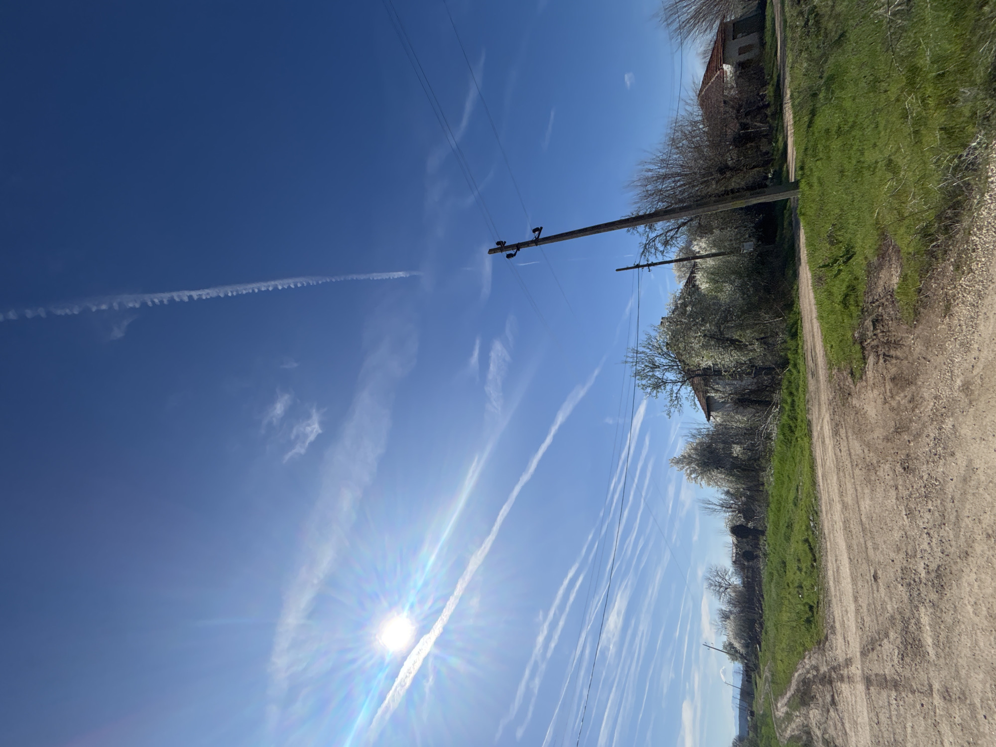 The image presents a dirt road in the village of Lehchevo, Bulgaria. Adjacent to the road is grassy terrain punctuated with trees and foliage. In the distance, a structure with a red roof is partially visible. Overhead, a clear blue sky is crisscrossed with contrails and power lines supported by wooden poles. The sun is shining brightly, creating a lens flare effect in the lower-left portion of the frame. The photograph is oriented in a vertical format.