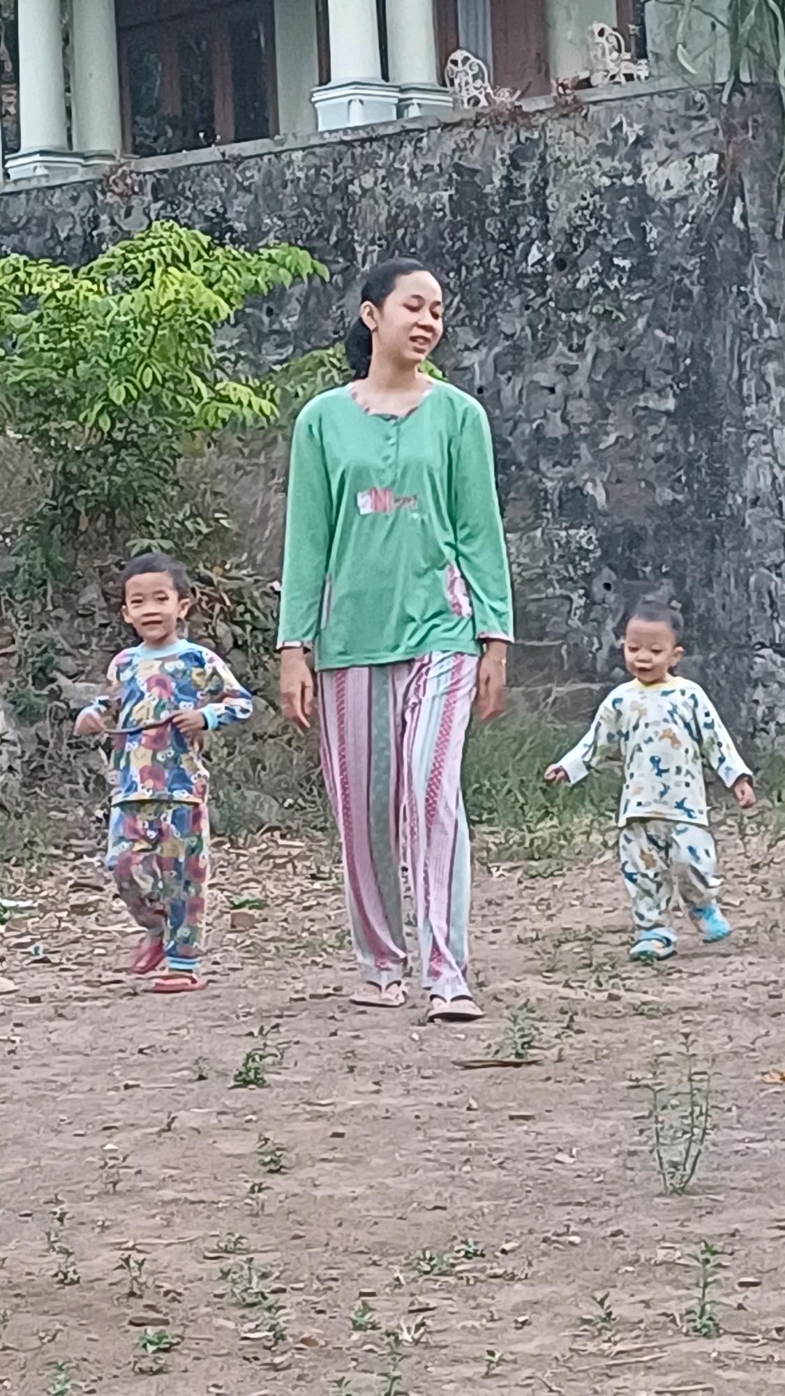 A mother and two young children are outdoors in the city of Wonokerso, Indonesia. The mother, wearing a green long-sleeved shirt and striped pants, walks slightly ahead of the two children. A boy on the left, wearing a multicolored patterned pajama set, looks towards the camera and holds a stick. A younger child on the right, in a light-colored pajama set with blue shoes, walks with arms outstretched. The ground is dirt with sparse vegetation. Behind them is a stone wall and part of a building with white pillars.