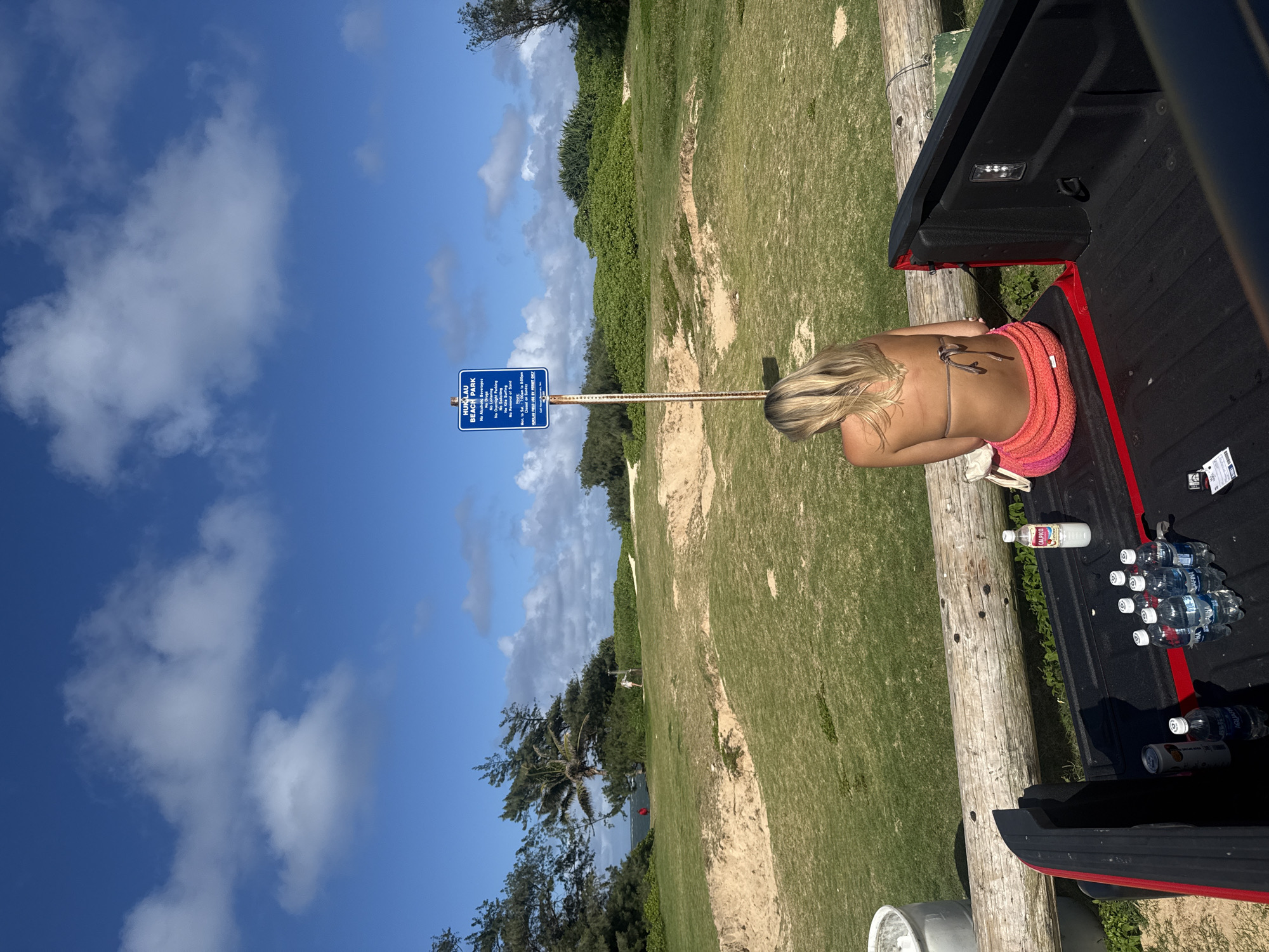 The image depicts a woman in a bikini and wrap sitting in the back of a pickup truck at Hukilau Beach Park in Laie, United States. The woman has blonde hair and is facing away from the camera towards a blue park sign indicating "HUKLAU BEACH PARK". The sign lists regulations prohibiting alcoholic beverages, drugs, loitering, overnight parking, and soliciting. The pickup truck bed contains several water bottles and a drink. The environment includes grass, trees, sand, and a blue sky with clouds.