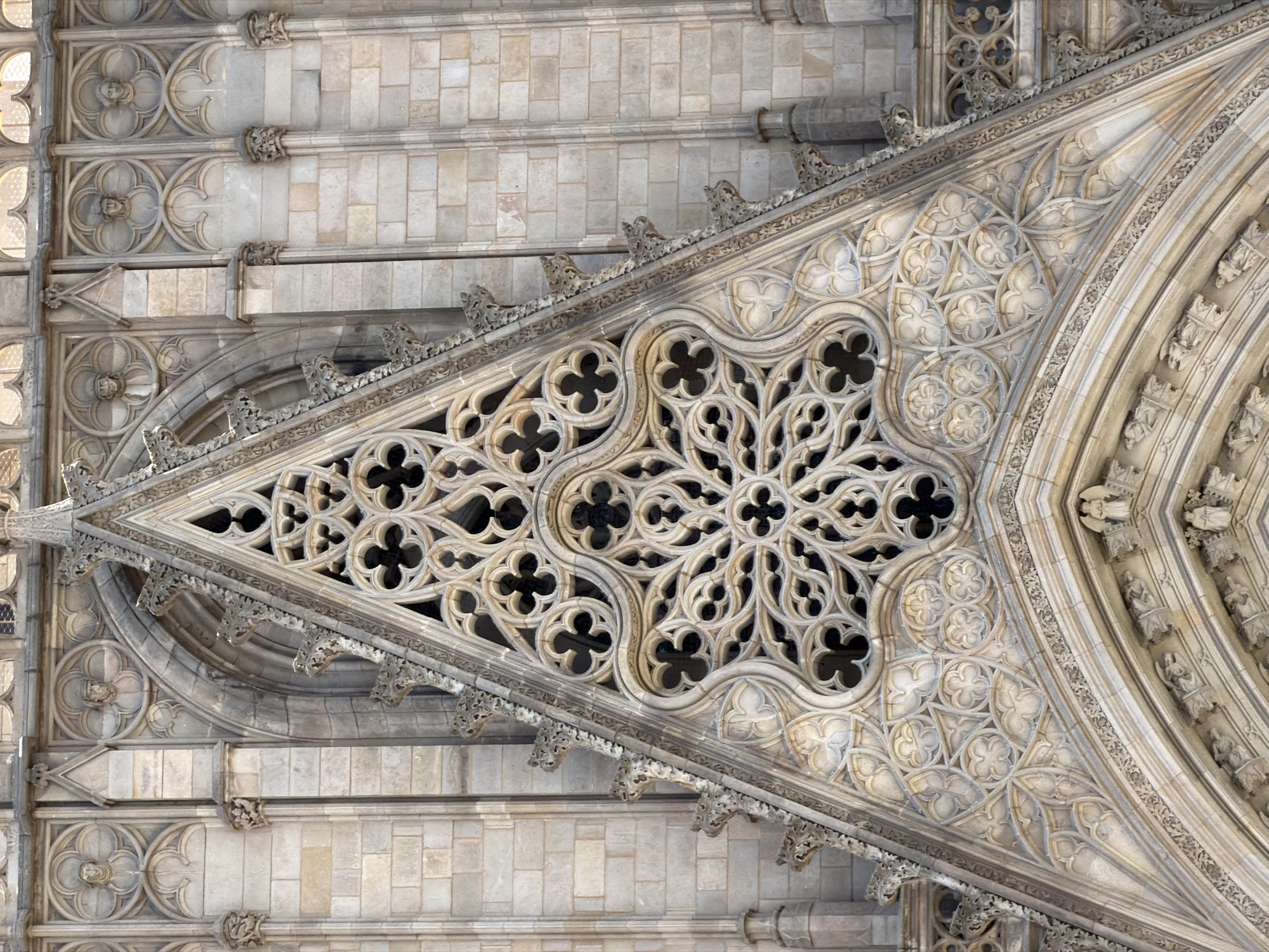 This media file is an overhead, close-up view of the intricate stone carvings on the exterior of a Gothic-style building, likely a cathedral, in Barcelona, Spain. The subjects are architectural elements, specifically ornate stonework and sculptures. The scene is a detailed examination of the building's facade, highlighting the craftsmanship involved.
The activity taking place is not depicted, as the image focuses on static architectural features. Notable details include the detailed tracery within a triangular pediment, featuring quatrefoil and trefoil motifs. The stone appears aged and weathered, suggesting it is an old structure. Small, carved figures, possibly statues or gargoyles, are visible along the decorative edges. The lighting suggests it is daytime, with sunlight casting shadows that accentuate the depth and texture of the carvings. The use of light and shadow adds a dramatic effect to the ornate details.
There is no visible text in the image. The overall impression is one of historical significance and artistic mastery in Gothic architecture. The setting is clearly an outdoor architectural landmark in Barcelona.