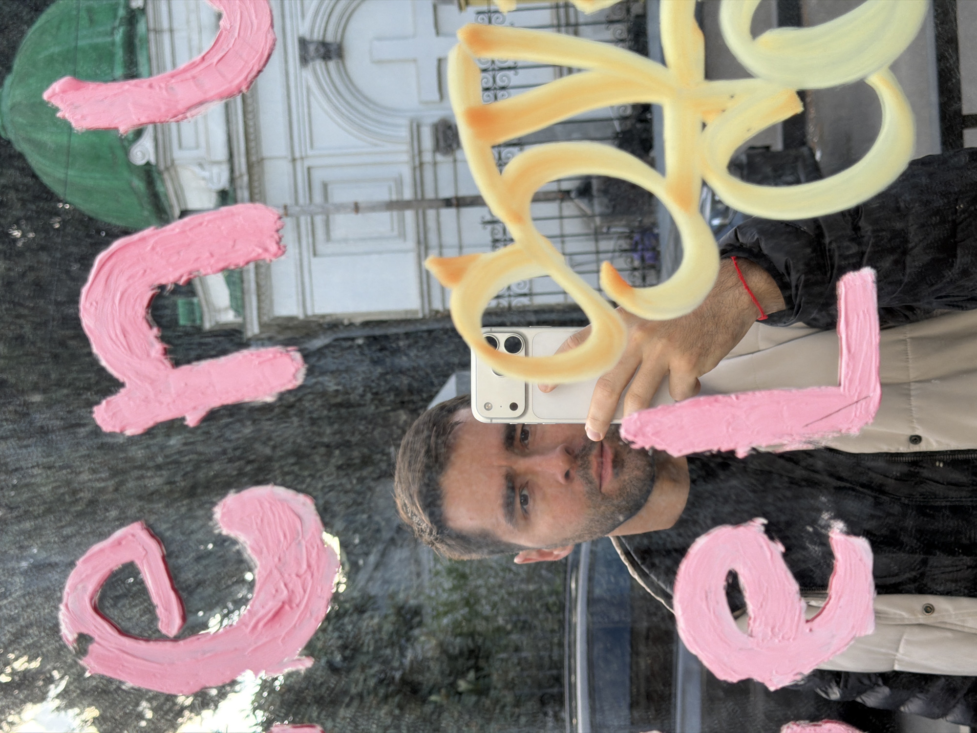 A man is taking a selfie in a reflective surface, which is smeared with pink and yellow paint in swirling and letter-like shapes. The reflection shows part of a cemetery gate. The cemetery is Luki Medea cemetery in Tbilisi, Georgia. The man has short brown hair, a beard, and is wearing a black jacket with a light-colored lining. He is holding a white phone with multiple cameras. The background shows glimpses of greenery and a white building with a rounded top and a cross. The photograph is taken at an angle, with the subjects appearing tilted.