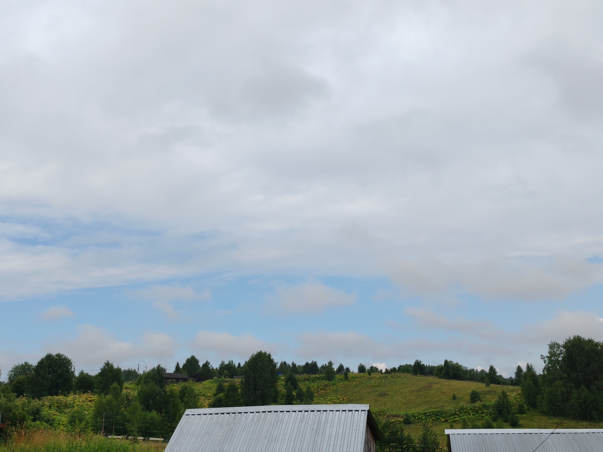 The image depicts a rural landscape under a cloudy sky in Чухлэм, Russia. In the foreground, the corrugated metal roof of a building is visible. Behind it, a green, rolling hillside is dotted with various trees and some vegetation. Several small, dark structures, likely houses or sheds, are nestled amongst the trees on the hillside. The sky is predominantly overcast with patches of blue sky showing through in the middle ground. The overall impression is one of a quiet, natural setting in the Russian countryside.