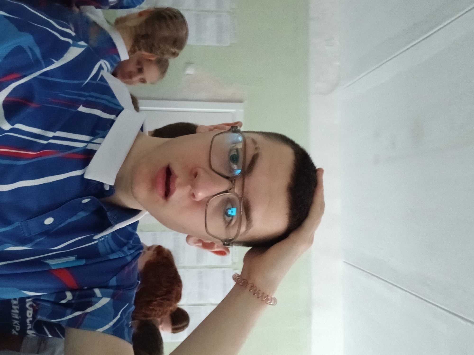 A young adult male with short dark hair and glasses is in the foreground, looking directly at the camera with a slightly surprised expression. He is wearing a blue polo shirt with white and red stripes and a white collar. His right hand is resting on his head, and a spiral hair tie is on his wrist.
In the background, there are several other people, some slightly blurred. It seems to be an indoor setting with light green walls and what appear to be notices or papers posted on them.
Given the information "город Березники, Россия" (Berezniki city, Russia) visible on the polo shirt, the location is likely Berezniki, Russia.