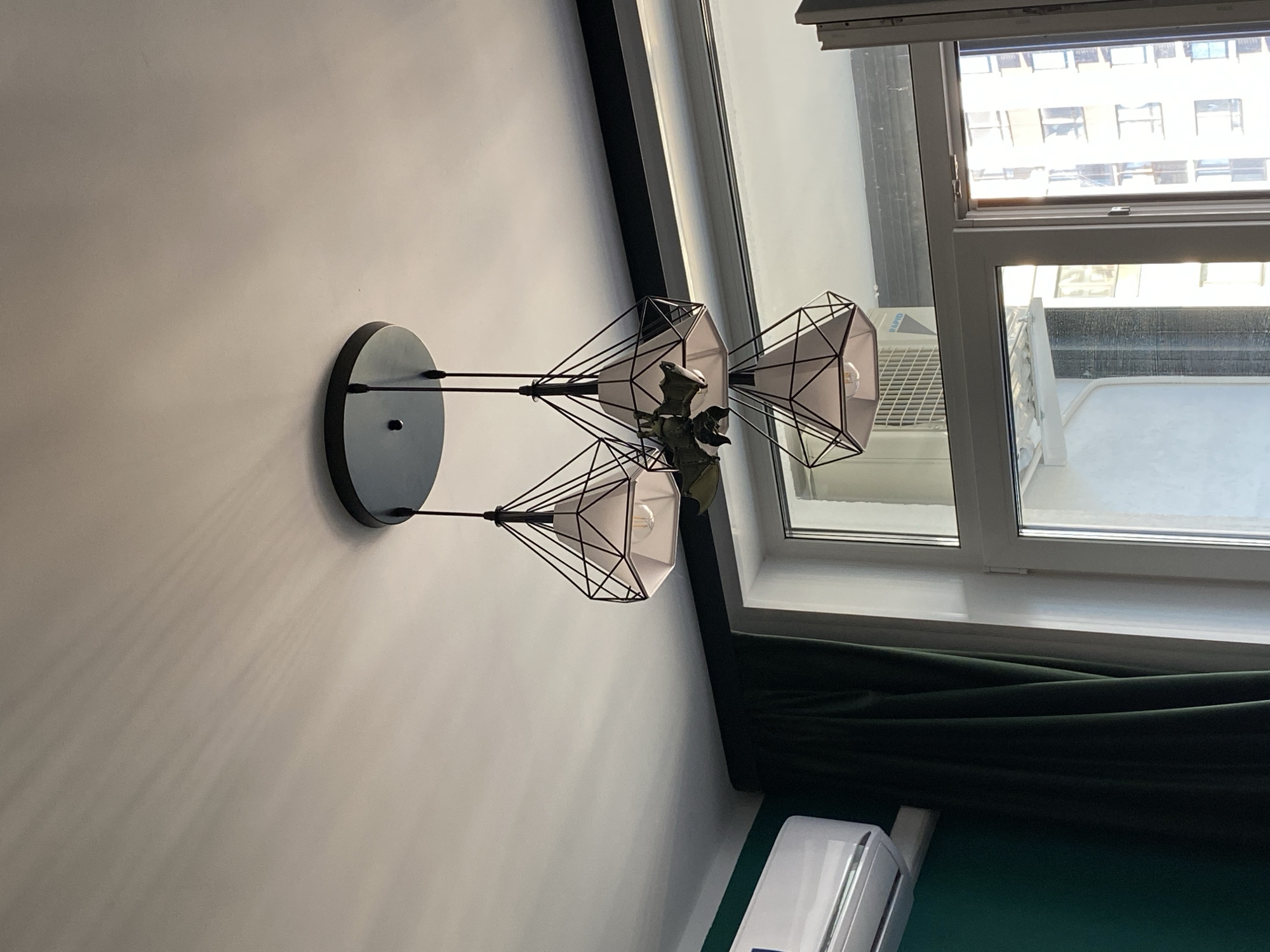 The image shows a ceiling lamp with three diamond-shaped shades. The lamp is made of black metal and has a circular black base attached to the ceiling. Each shade contains a light bulb. A decorative artificial plant is attached to the central part of the lamp. To the left of the lamp, part of a white air conditioning unit is visible on a dark green wall. A dark green curtain hangs to the right of the air conditioning unit. A window with white framing is visible in the lower right portion of the image, looking out onto a building. The scene appears to be indoors. The image does not contain any people or specific identifiable landmarks of Yekaterinburg, Russia.