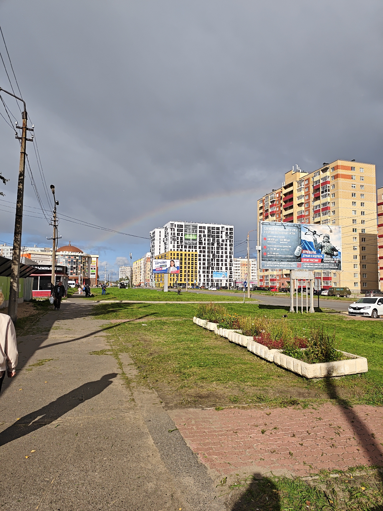 The image captures a street scene with a rainbow arching across a cloudy sky over a city. In the foreground, a paved sidewalk runs alongside a grassy area with a long, raised planter bed containing flowers and vegetation. A person's shadow stretches across the sidewalk. To the left, a utility pole with overhead wires stands near a small structure with an awning. Several buildings are visible in the midground and background, including modern apartment complexes with balconies and windows, and a building with a domed roof. Billboards are also present, one featuring a recruitment advertisement for military service and another with a portrait of a woman. Cars are parked along the street and on a roadway in the distance. The environment appears to be an urban residential area on a partly sunny day with some clouds.