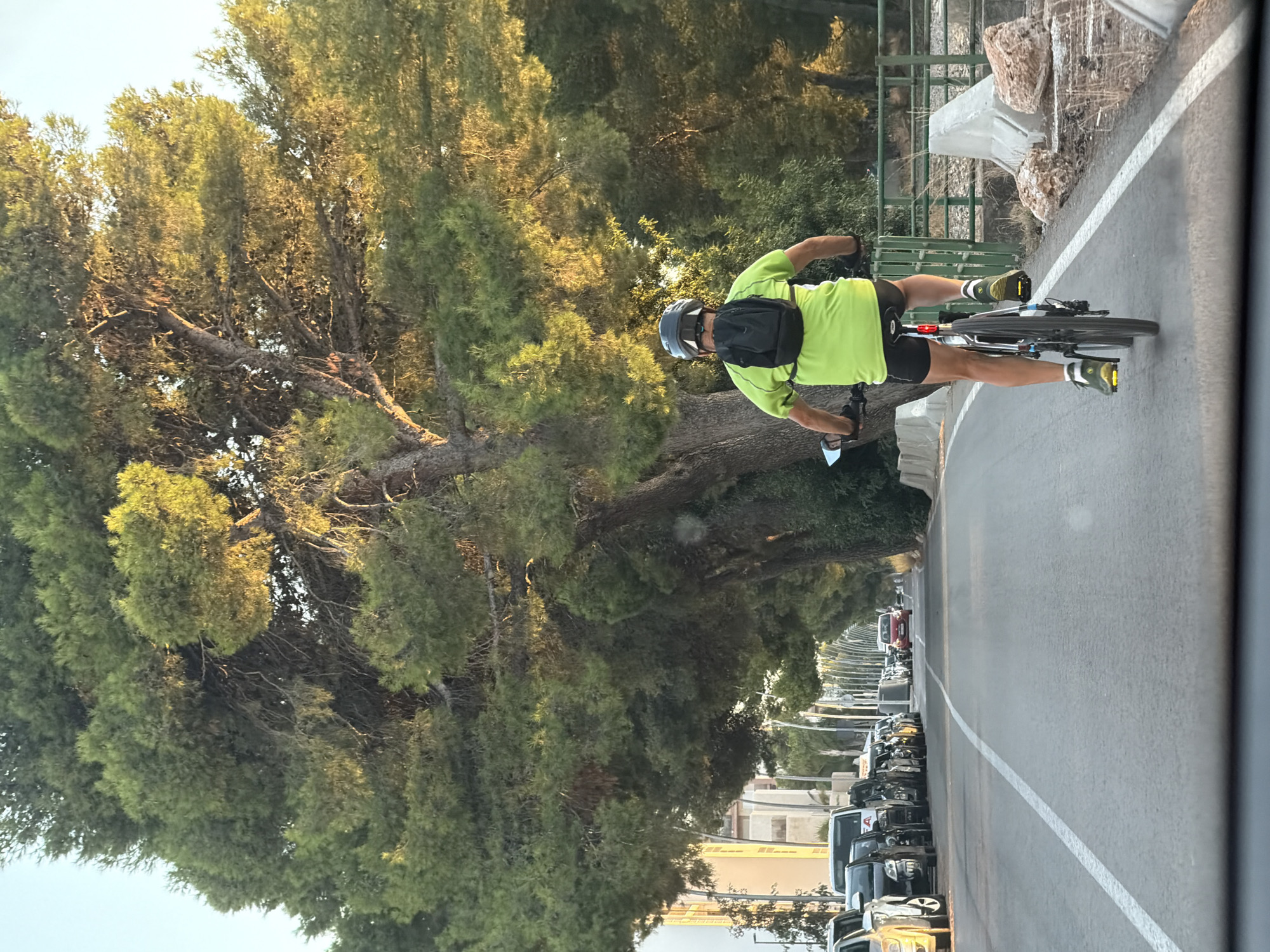 The image shows a man cycling on a road in Mont-roig del Camp, Spain. He is wearing a bright green t-shirt, black shorts, a helmet, and a backpack. He is riding a mountain bike with a black frame and silver handlebars. The road is paved and has a white line marking the lane. On the left side of the road, there are several parked cars and buildings. On the right side of the road, there is a fence and some rocks. The background is filled with lush green trees, which are casting long shadows across the road, suggesting it is late afternoon or early evening. The sky is clear and blue. The scene appears to be a typical urban or suburban street with moderate traffic and ample greenery.