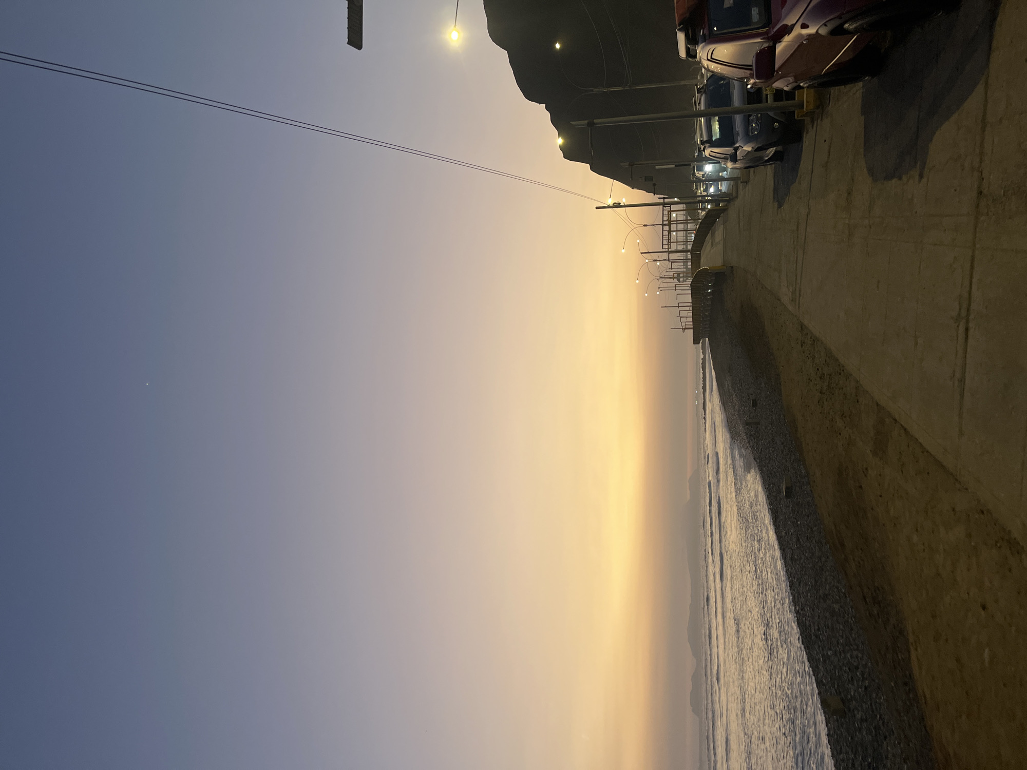 The image depicts a sunset scene along the Miraflores coastline in Peru. A paved walkway runs parallel to the ocean, which features gentle waves breaking on a pebbly shore. On the right side of the walkway, several parked vehicles, including SUVs and a red vehicle in the foreground, are visible. Streetlights and power lines are also present, casting a warm glow in the fading light. A rocky incline rises behind the walkway and to the right, characteristic of the Miraflores Malecón. The sky transitions from a pale purple at the top to a vibrant orange and yellow near the horizon, with distant islands silhouetted against the colorful sky. There are no people visible in the image.