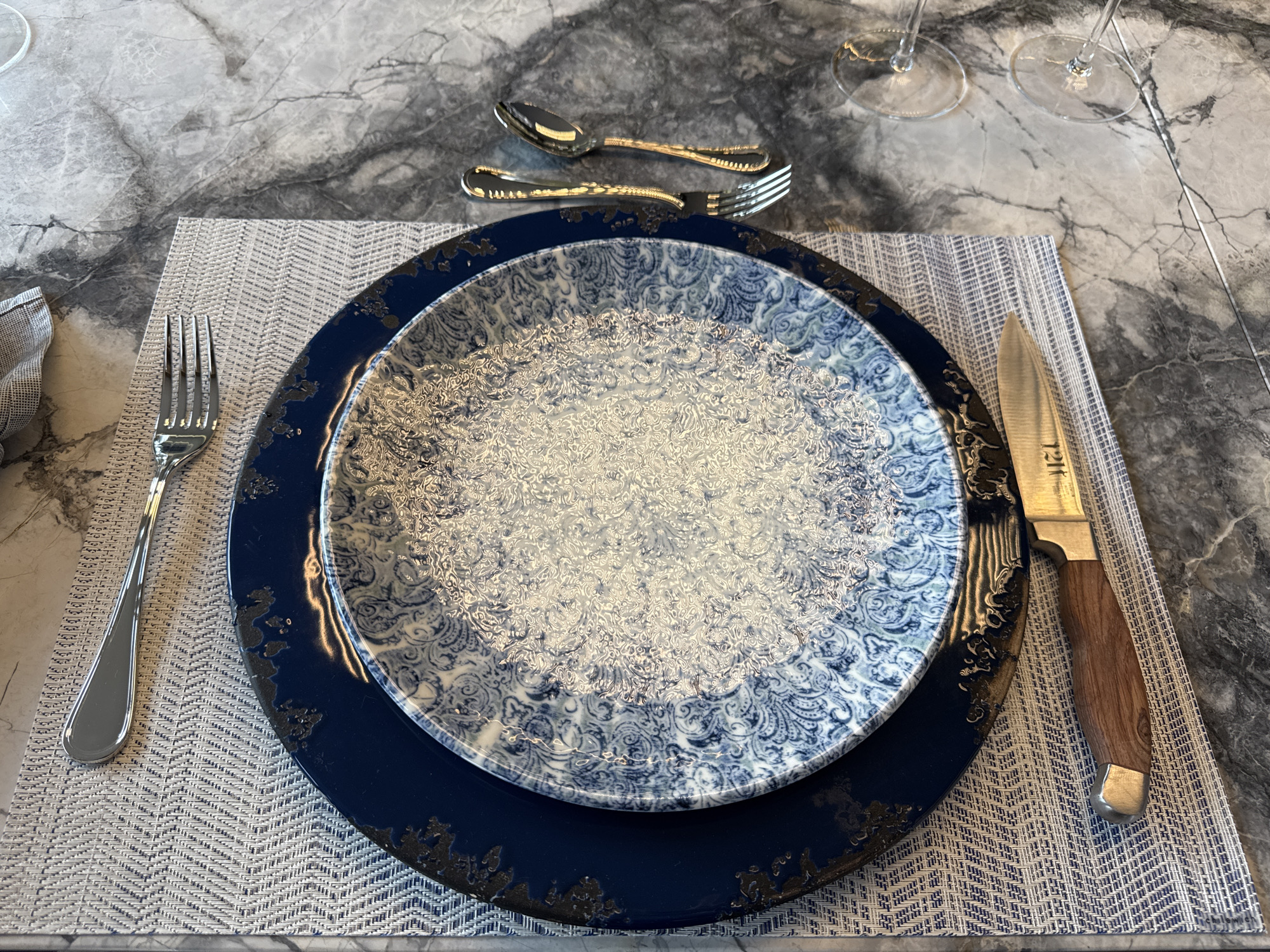 A place setting is arranged on a gray-and-white placemat. A silver fork is placed to the left of the placemat, and a knife with a wooden handle is placed to the right. Two stacked plates, one navy blue with distressed edges and a smaller white plate with a blue floral pattern, occupy the center of the placemat. Above the plates are a silver spoon and fork. In the background are two wine glasses. The table is made of gray marble. The image was likely taken indoors. There is no indication of location in Mexico City, Mexico.