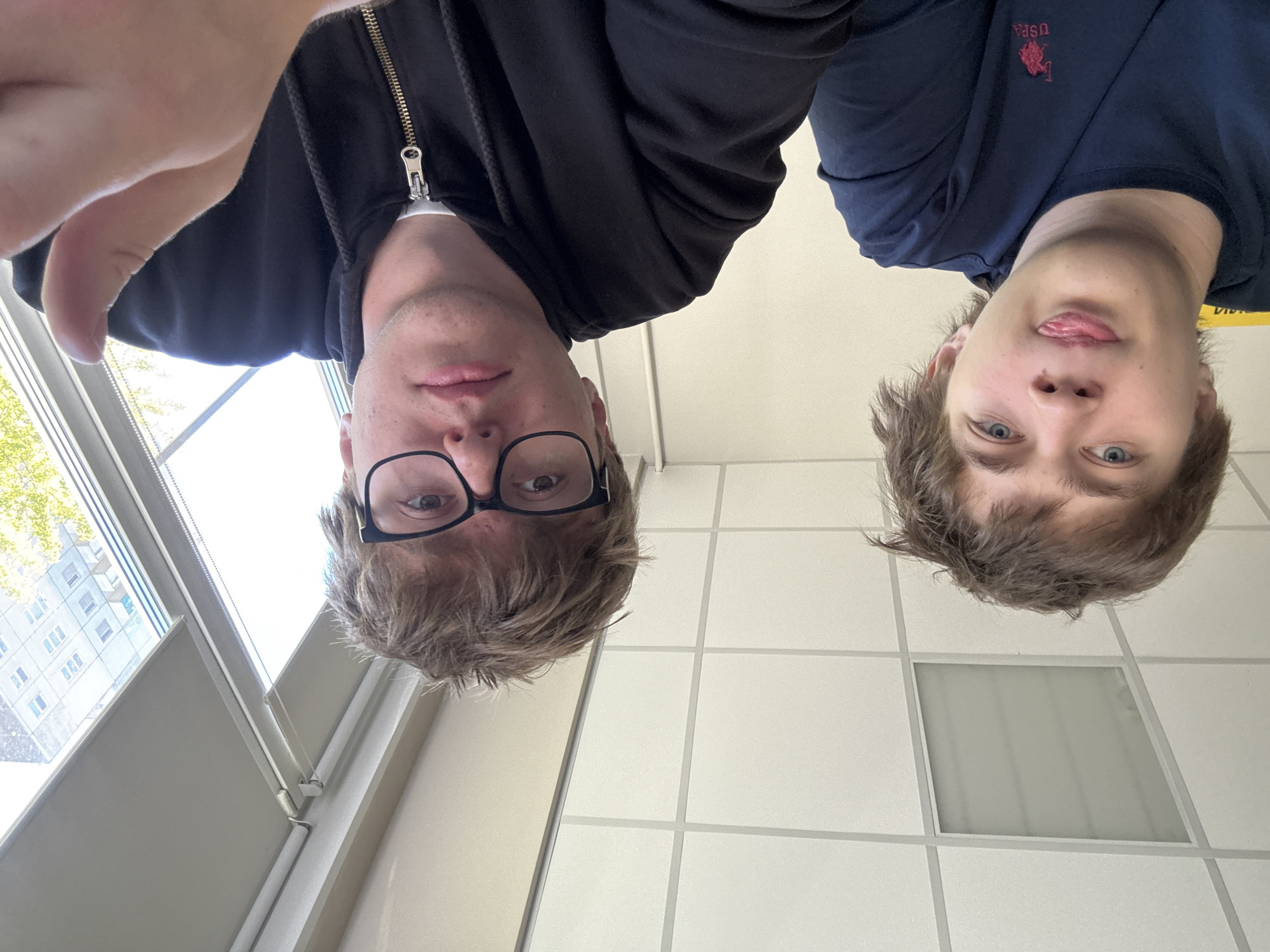 Two young men are positioned with their faces oriented towards the camera, which is held at an elevated angle, creating a perspective looking down at them. The man on the left wears glasses and a dark-colored hooded jacket. The man on the right is wearing a navy blue polo shirt with a logo on the upper left side. The background consists of a window with blinds on the left and a tiled ceiling on the right. A building can be seen through the window, suggesting an indoor location. It is impossible to determine the location in the city of Krasnoyarsk, Russia from this image.