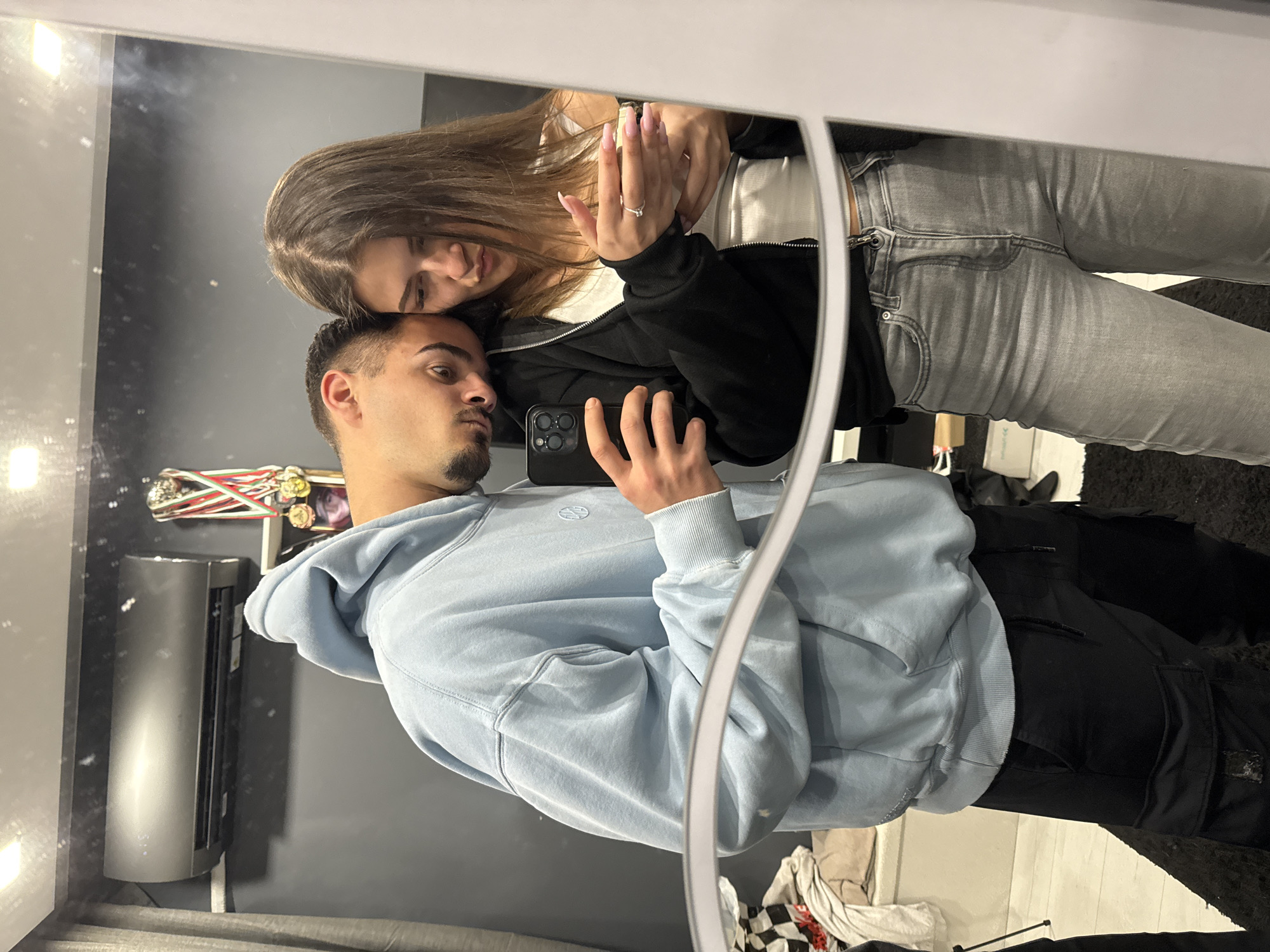 A couple is taking a selfie in a mirror. The man is wearing a light blue hoodie and black pants. The woman is wearing a white top, a black jacket, and grey jeans. She has long brown hair and is leaning her head on the man's head. The man is holding a phone with both hands to take the picture. The background includes a dark grey wall with an air conditioning unit, a shelf with medals and ribbons, and a black rug. The location appears to be indoors, possibly in an apartment or home. There is no information in the image to indicate that it is in Sofia, Bulgaria.
