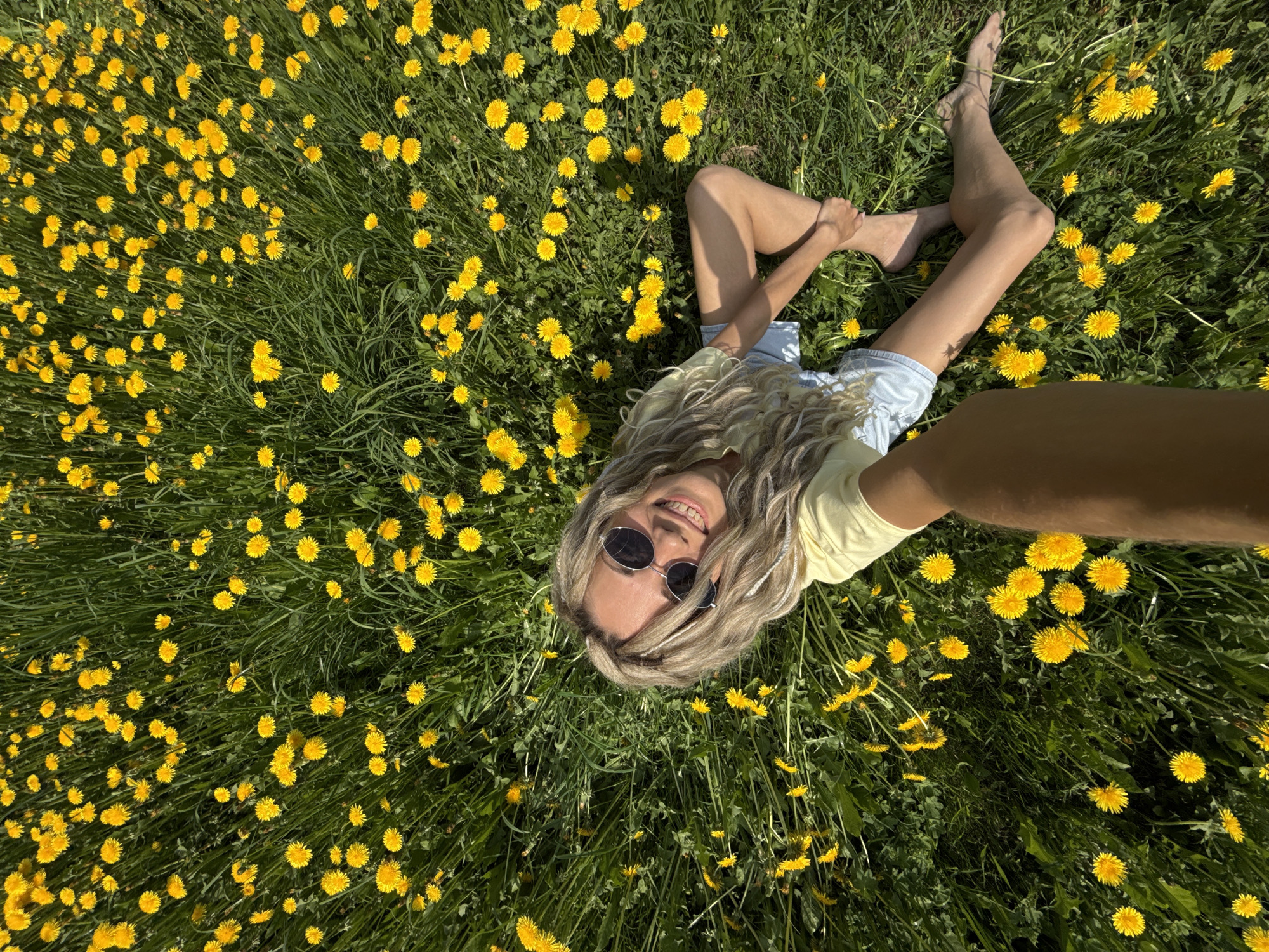 A woman lies in a field of yellow dandelions. She is wearing sunglasses, a yellow shirt, and light blue shorts. Her feet are bare. One arm is extended towards the camera as if taking a selfie. The location is in a field of dandelions in the city of Unknown, Unknown.