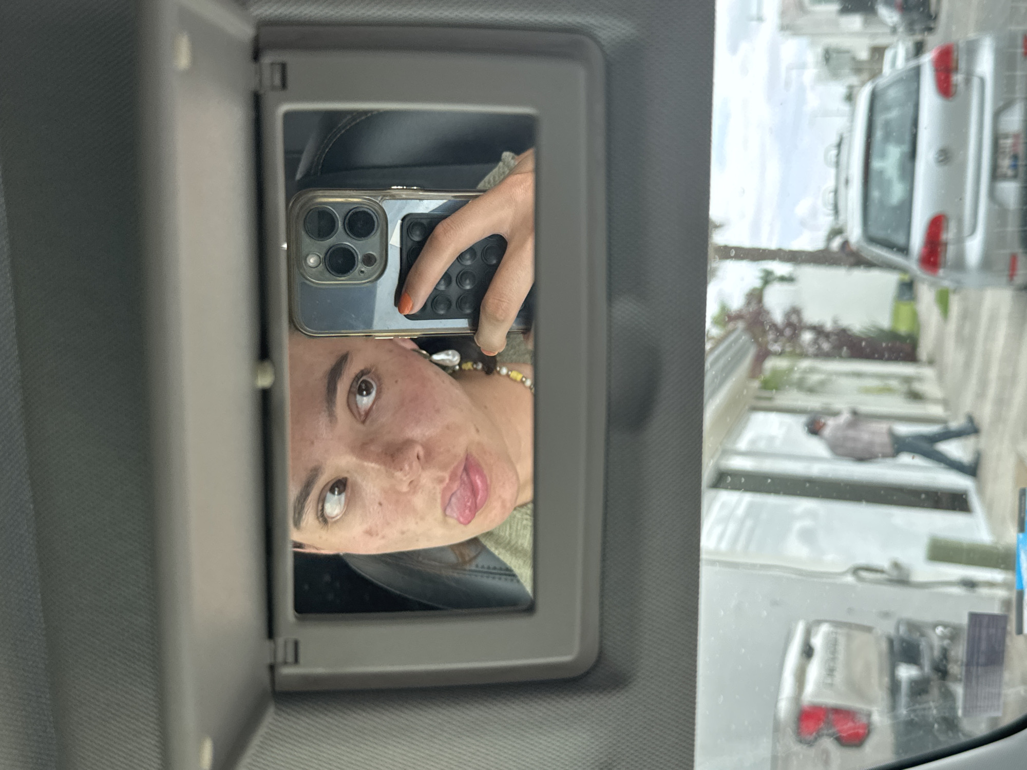 The image is a selfie taken in a car's visor mirror. A young woman with freckles is sticking out her tongue and looking to the left, holding a phone in her right hand. She is wearing a green shirt and a beaded necklace. A pearl earring is visible on her left ear. The phone has a clear case with a pop socket attached. The background shows the interior of the car and a view through the windshield of a street in San Bernardino Tlaxcalancingo, Mexico. A silver car is parked on the street, and a person in a light-colored shirt and jeans is walking away from a building. The street is lined with houses.