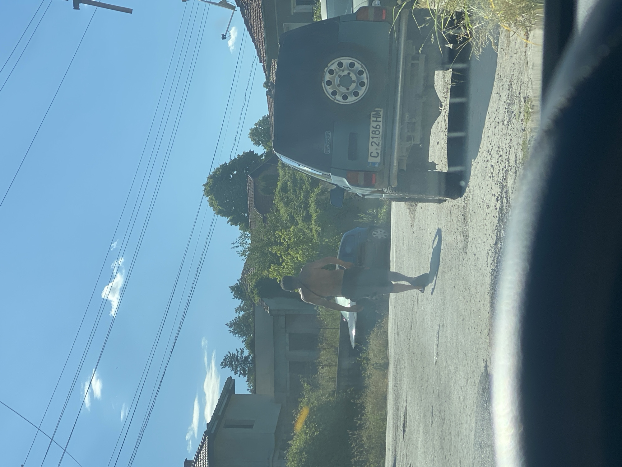 The image depicts a street scene in the city of Sadovets, Bulgaria, on a sunny day. A man, shirtless and wearing dark shorts and sandals, is walking away from the camera on a paved road. To the right of the man, a dark green SUV is parked, facing away from the camera, with a spare tire mounted on the back. The license plate of the SUV reads "C. 2186 HM". Behind the man and the SUV, several buildings with tiled roofs are visible, interspersed with lush green trees and vegetation. Overhead, power lines stretch across the clear blue sky, with a few wispy clouds present. The foreground is dominated by a dark, out-of-focus area, suggesting the photo was taken from inside a vehicle.
