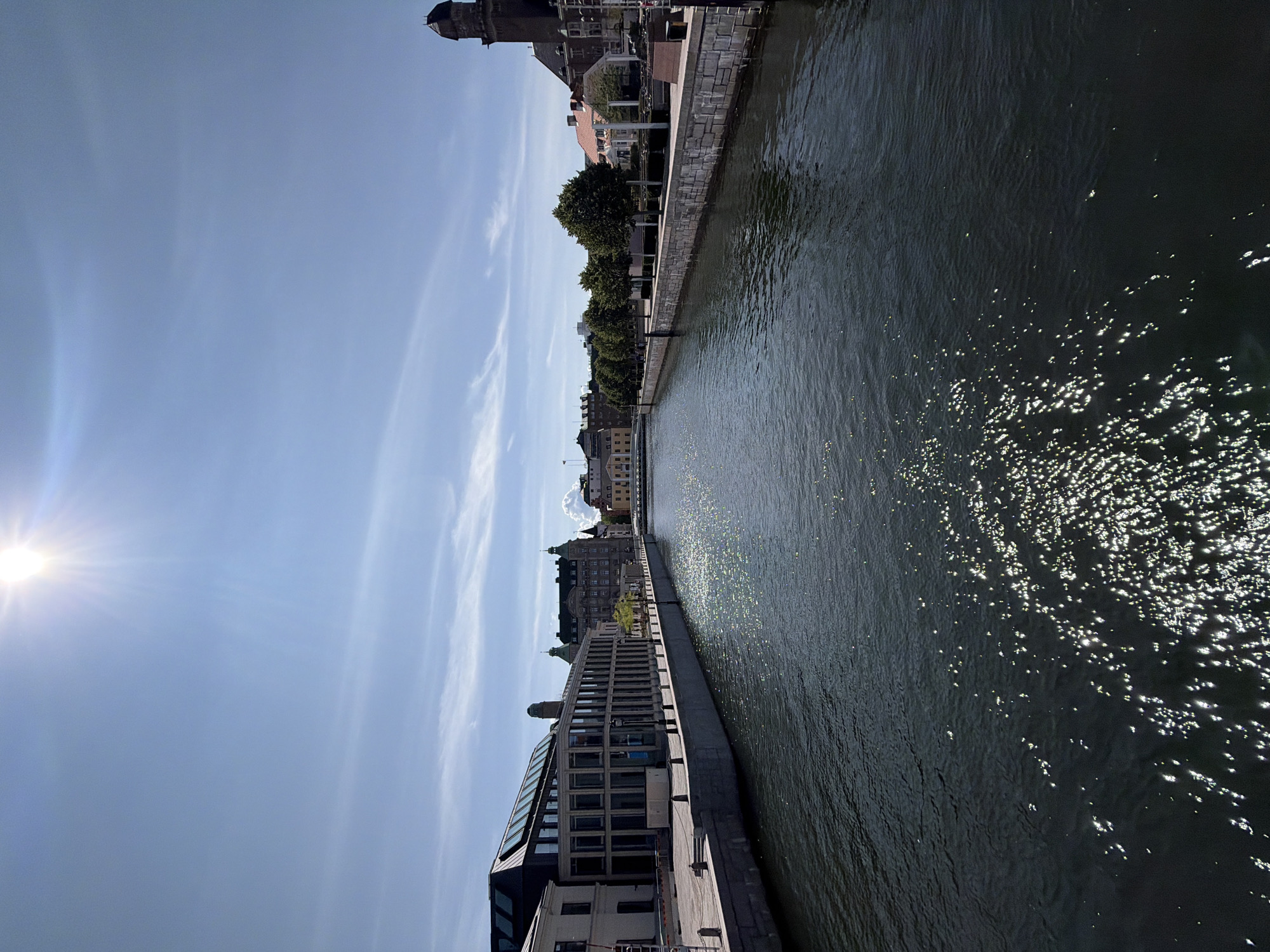 The image shows a view of a canal in Malmö, Sweden, under a bright sunny sky with some wispy clouds. The canal is wide and dark, with sunlight glinting off the surface. On either side of the canal are stone embankments, with buildings lining the banks.
On the left side of the canal, there is a modern building with a glass facade and a dark, angular roof structure. Further down this side, there are more traditional buildings with lighter colored facades.
On the right side of the canal, there are trees planted along the embankment, behind which are buildings with red-tiled roofs and a tall, dark-colored tower. The architecture appears to be a mix of historical and modern styles.
The scene is devoid of any visible people, animals, or vehicles, suggesting a peaceful and tranquil urban environment. The sun is high in the sky, indicating it is likely midday. The overall impression is one of a well-maintained cityscape with a prominent waterway.