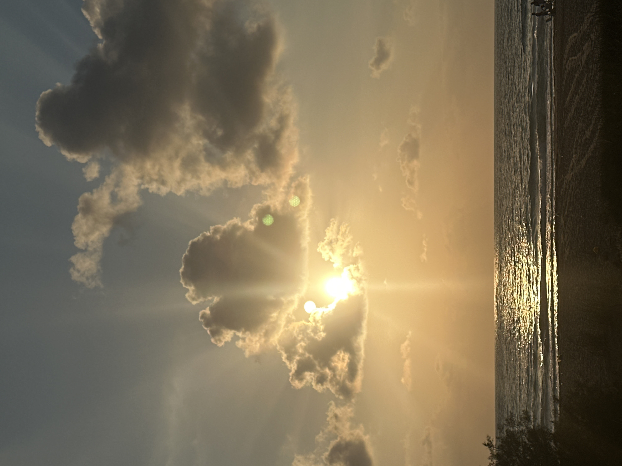 The image shows a beach scene during sunset, likely in the city of Mate Asher Regional Council, Israel. The sun is partially obscured by clouds, casting golden light and rays across the sky and the sea. The ocean surface is illuminated with a shimmering reflection of the sun, with gentle waves rolling towards the sandy shore. In the foreground, the beach is dark, with some vegetation on the left side. A few people are visible on the sand in the distance, appearing as small figures silhouetted against the bright horizon. The sky is filled with a mix of clouds, some dark and heavy, others lighter and illuminated by the setting sun.