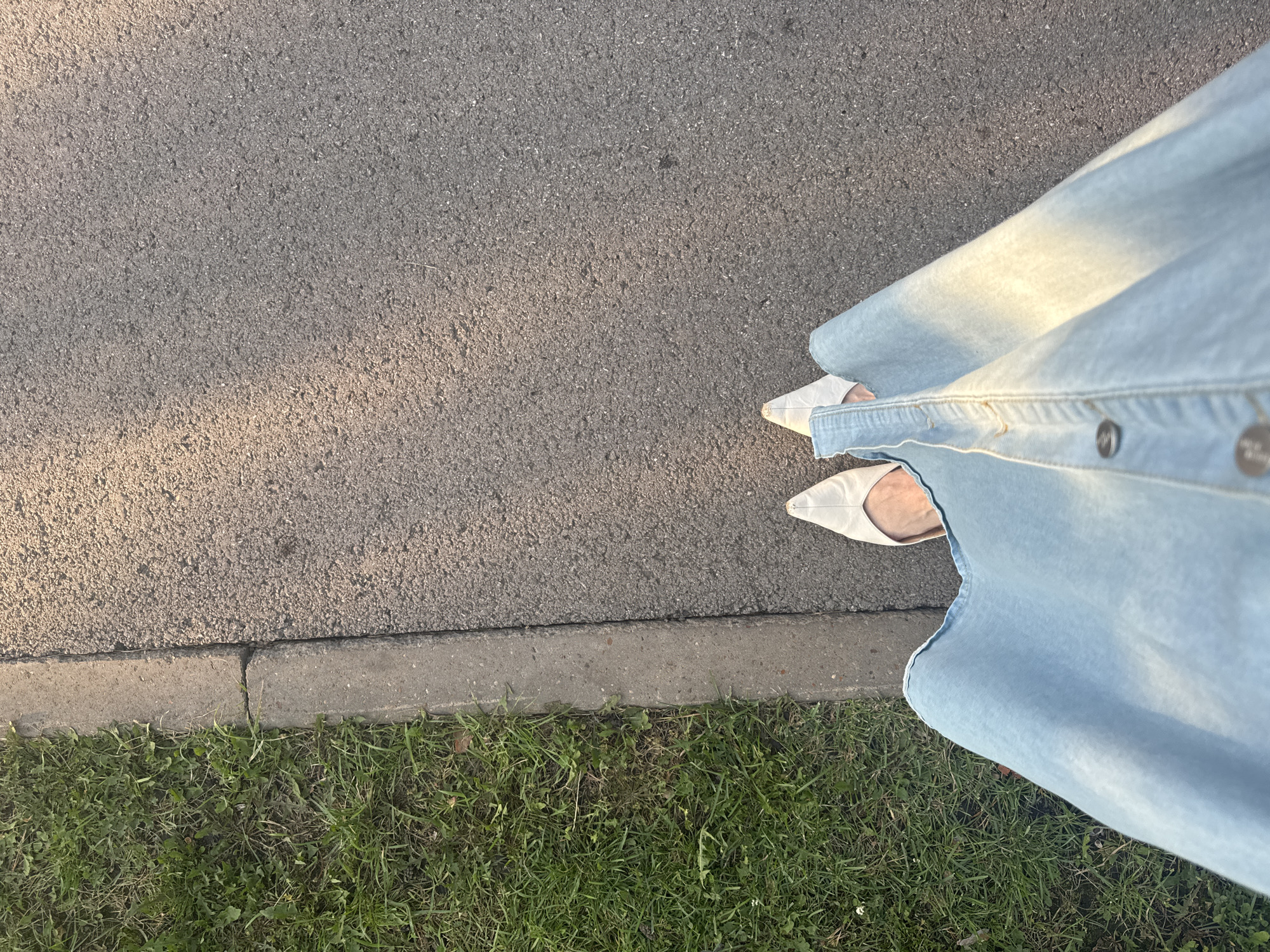 The image shows a person's perspective from above, looking down at their feet and legs. They are standing on an asphalt surface next to a grass area separated by a concrete curb. The person is wearing light-colored, pointed-toe shoes and a light blue denim skirt or dress. The lighting suggests it is daytime, possibly late afternoon due to the warm light casting shadows. The scene depicts a typical urban or suburban setting, possibly a park path or sidewalk. The location in Ufa, Russia cannot be determined from the visual information alone.