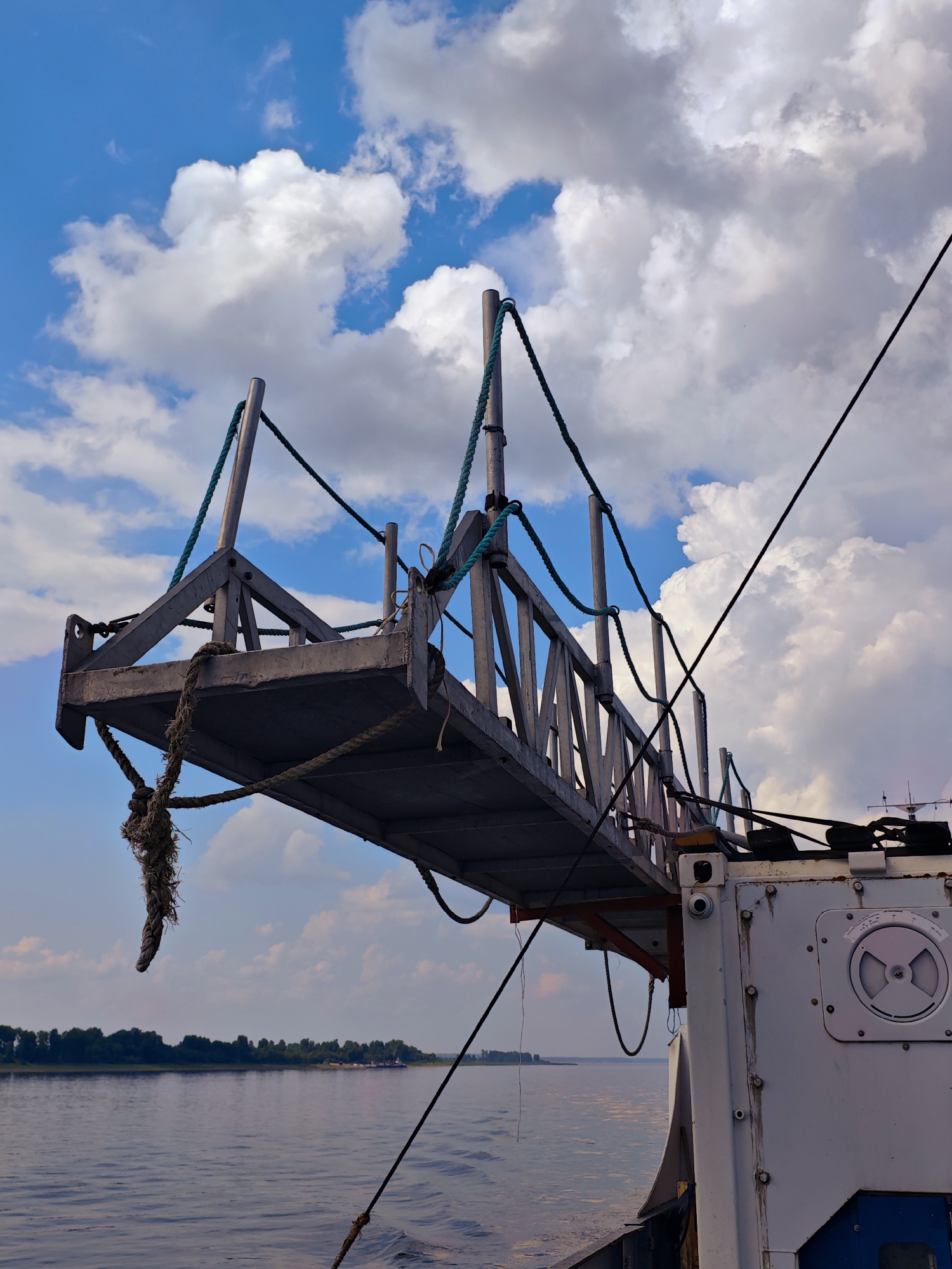 A metal gangplank extends from a vessel onto a calm body of water under a partly cloudy blue sky. The gangplank is supported by ropes and metal struts and appears weathered. The side of the vessel is white with visible bolts and a circular gauge. In the distance, a tree-lined shore is visible, and on the water, a barge is present. The overall scene suggests a river or harbor environment. The location is Erkalovo, Russia.