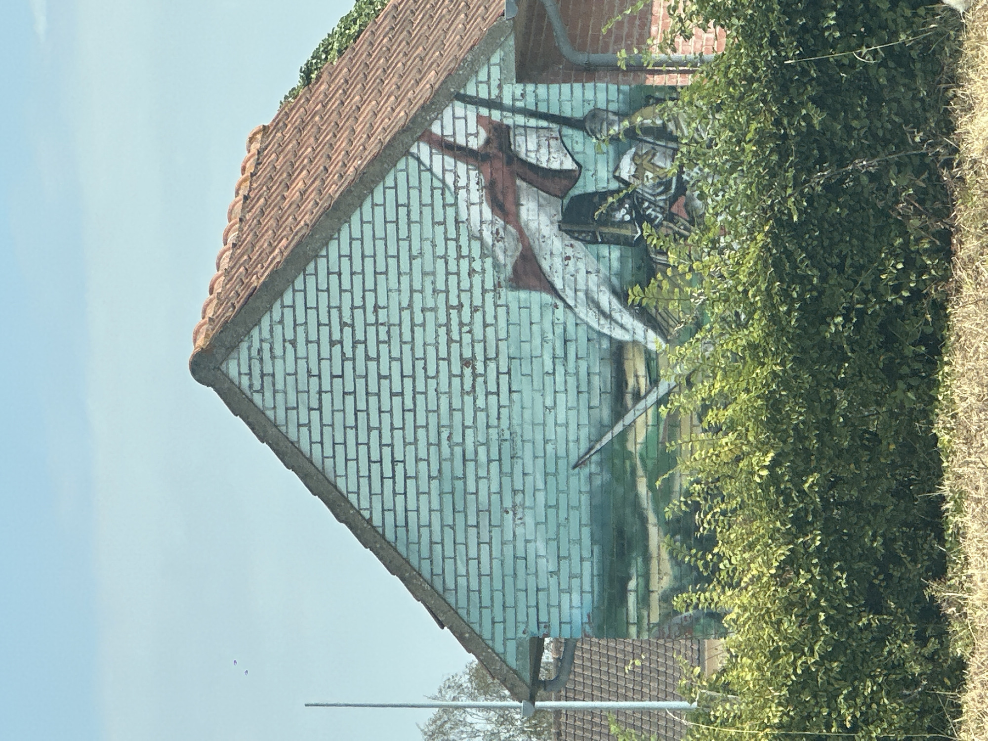 The image depicts the gable end of a brick building, painted with a mural. The mural features a figure in armor, possibly a knight, holding a flag with red and white markings. The background of the mural appears to be a landscape. The building has a tiled roof and is partially obscured by green foliage in the foreground. The sky is blue and clear. A thin vertical pole is visible to the left of the building. The mural is located on a building in Conchil-le-Temple, France.