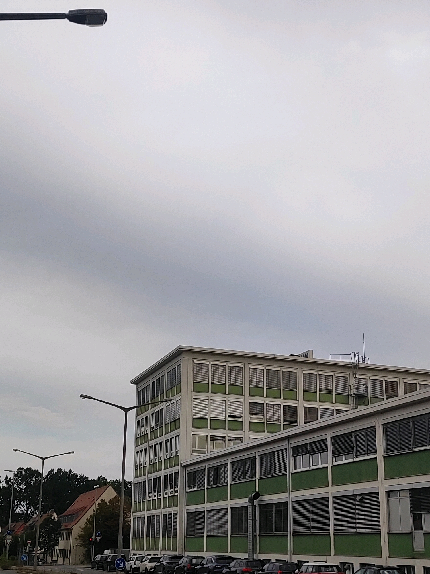 The image depicts a cloudy, overcast sky over an industrial or commercial area in Nuremberg, Germany. In the foreground, a tall, dark lamppost extends from the left side of the frame. To the right, a multi-story building with a distinctive green facade and numerous windows with blinds is prominent. The building appears to be an office or factory. Further to the left, another lamppost is visible, and a row of parked cars is situated in front of the building and along the street. Behind the building, several smaller houses with red roofs and trees are visible, suggesting a mix of urban and residential development. There are traffic signs and signals visible on the street. No people are clearly visible in the frame. The overall scene is one of a functional, daytime urban environment under a muted sky.