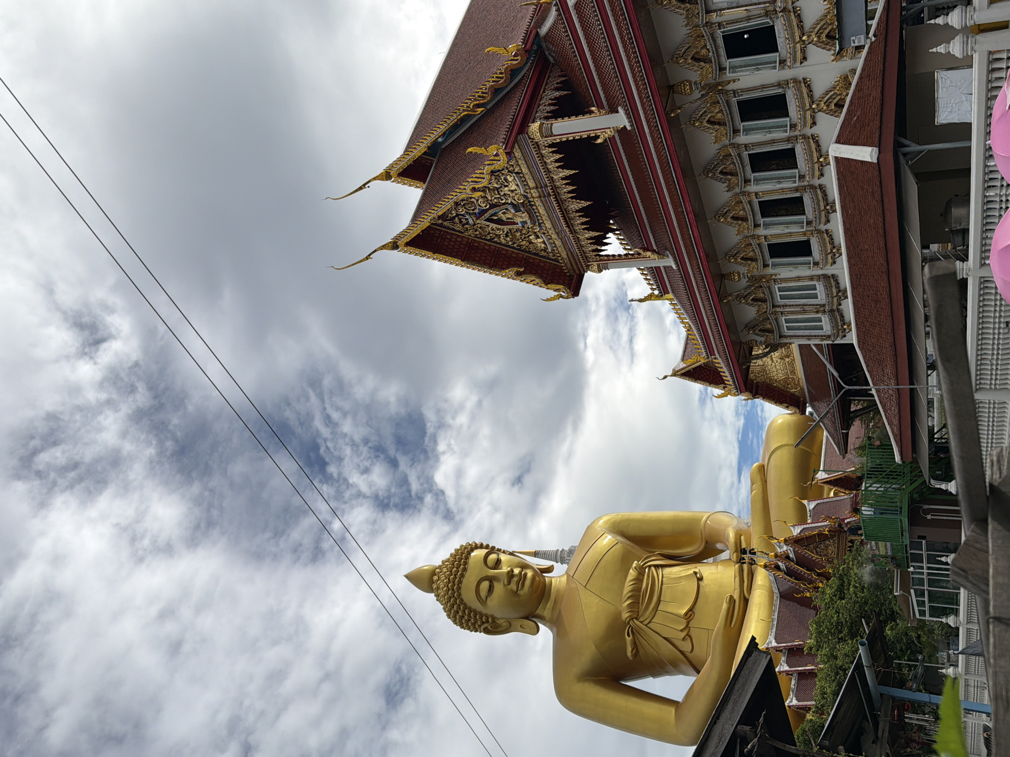 The image depicts a large, golden Buddha statue sitting in a meditation pose, seen from the chest up, with its face serene and eyes closed. To the right of the statue is a red-roofed temple building with ornate golden decorations on its eaves and facade. The sky is cloudy, with streaks of white clouds against a blue background, and two power lines are visible traversing the upper portion of the frame. Below the Buddha and temple, there are glimpses of other structures and lush green trees. In the foreground, a white railing is visible. The scene appears to be an outdoor view of a temple complex. This is the Wat Muang temple in Ang Thong province, near Bangkok, Thailand, which houses one of the world's largest Buddha statues.
