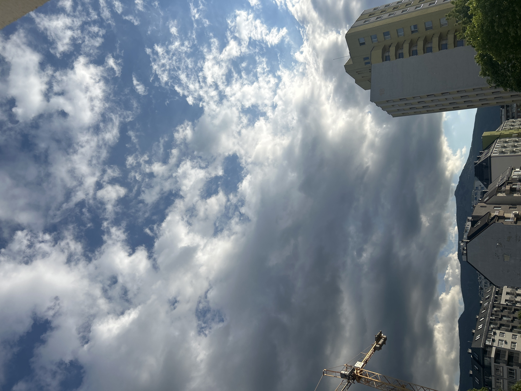 The image shows an upward view in Sofia, Bulgaria, dominated by a cloudy sky with patches of blue. A crane is visible in the lower right corner. Adjacent to the sky, buildings and a mountain range are partially visible. A tall, yellow building sits at the upper right corner.