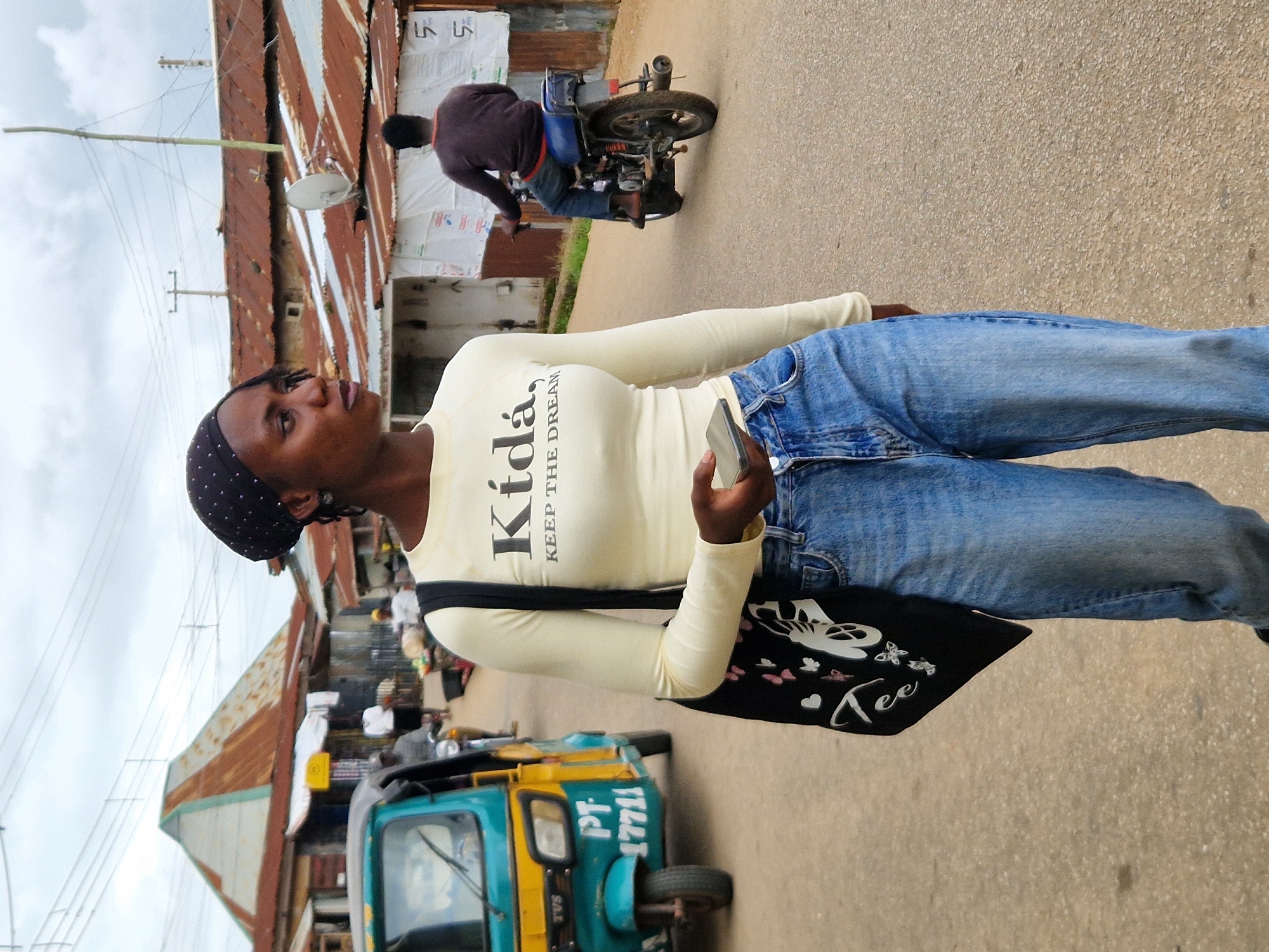 This is a street-level, medium shot photograph taken in the city of Jos, Nigeria. The image captures a young woman walking towards the viewer, holding a smartphone in her right hand. She is wearing a light-colored, long-sleeved shirt with the text "Kida, KEEP THE DREAM" printed on it in black, and light-wash denim jeans. She has a black headscarf on and a large black tote bag with white butterfly and butterfly wing designs slung over her left shoulder.
Behind her, a man on a motorcycle is riding away from the viewer, also moving down the paved street. Further in the background, several buildings with corrugated tin roofs line the street. One building on the left has a triangular, light-colored roof with rusted metal detailing. A turquoise and yellow three-wheeled auto-rickshaw, also known as a "Keke Napep" in Nigeria, is parked on the left side of the street. The sky is overcast, suggesting a cloudy day. The overall scene depicts a typical urban environment with elements of local transportation and commerce.