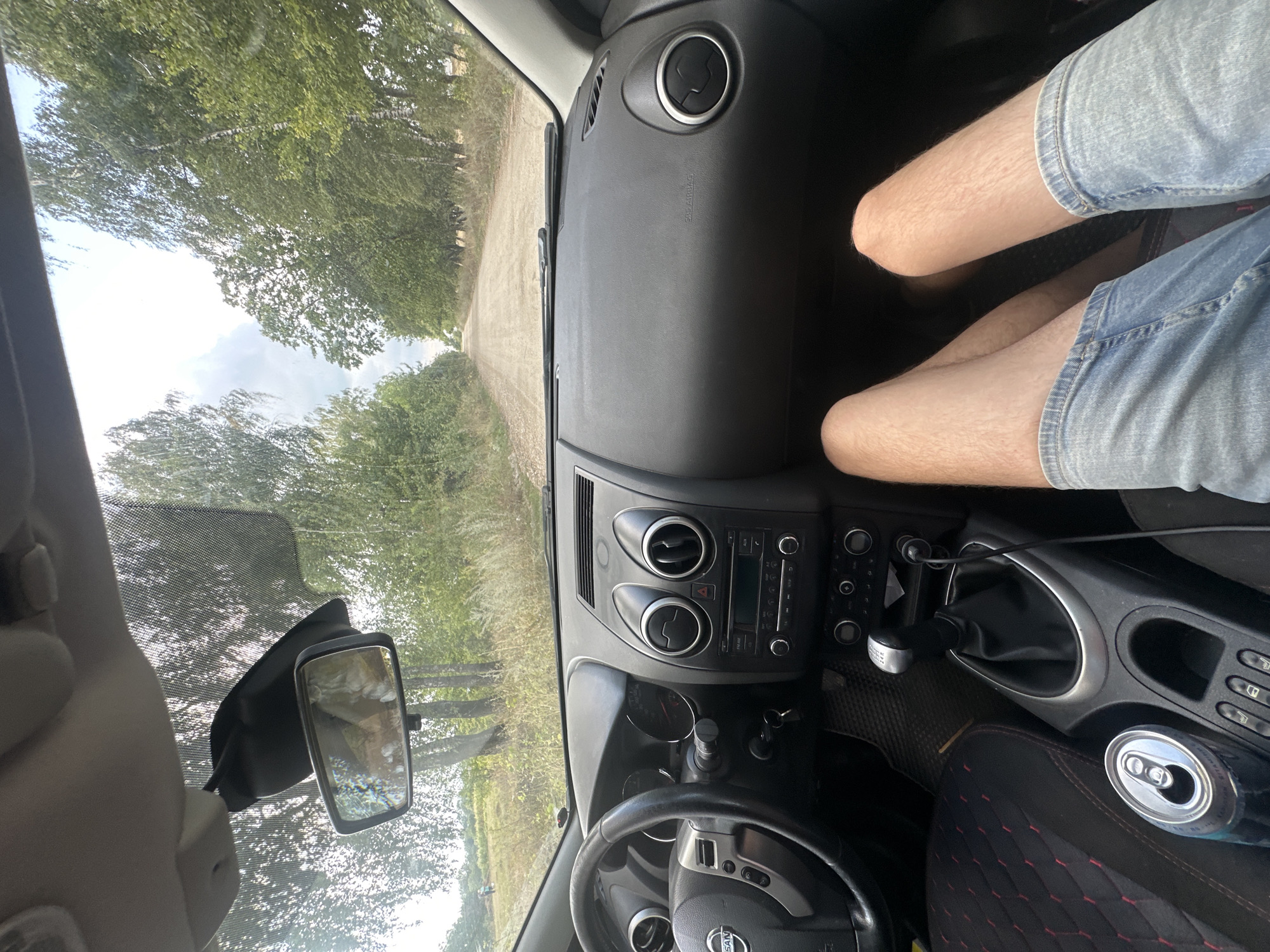 An interior, first-person perspective shot from a car shows a dirt road lined with trees extending into the distance. The dashboard is visible in the foreground, with the steering wheel on the left and the gear shift in the center. A person's legs, wearing denim shorts, are positioned in the lower right of the frame. A can of soda rests in a cup holder to the left of the gear shift. The scene appears to be set in a rural area, possibly on the outskirts of Tynna, Ukraine.