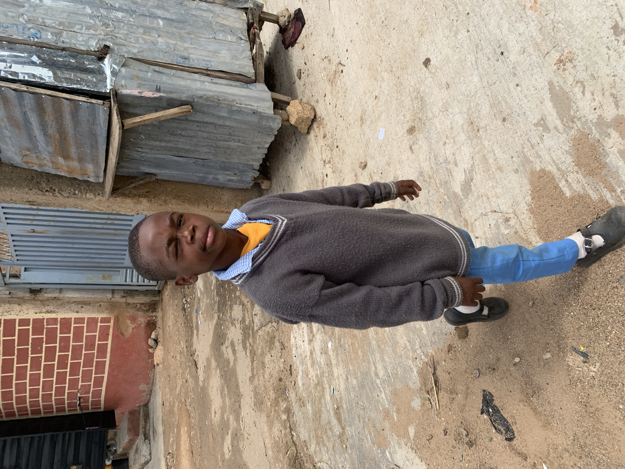 A child stands outdoors on a dirt and concrete surface. They are wearing a grey sweater, light blue pants, white socks, and black Mary Jane shoes. The child is looking directly at the camera. Behind the child is a structure made of corrugated metal, a building with brickwork visible, and a grey metal gate. The location is Jos, Nigeria.