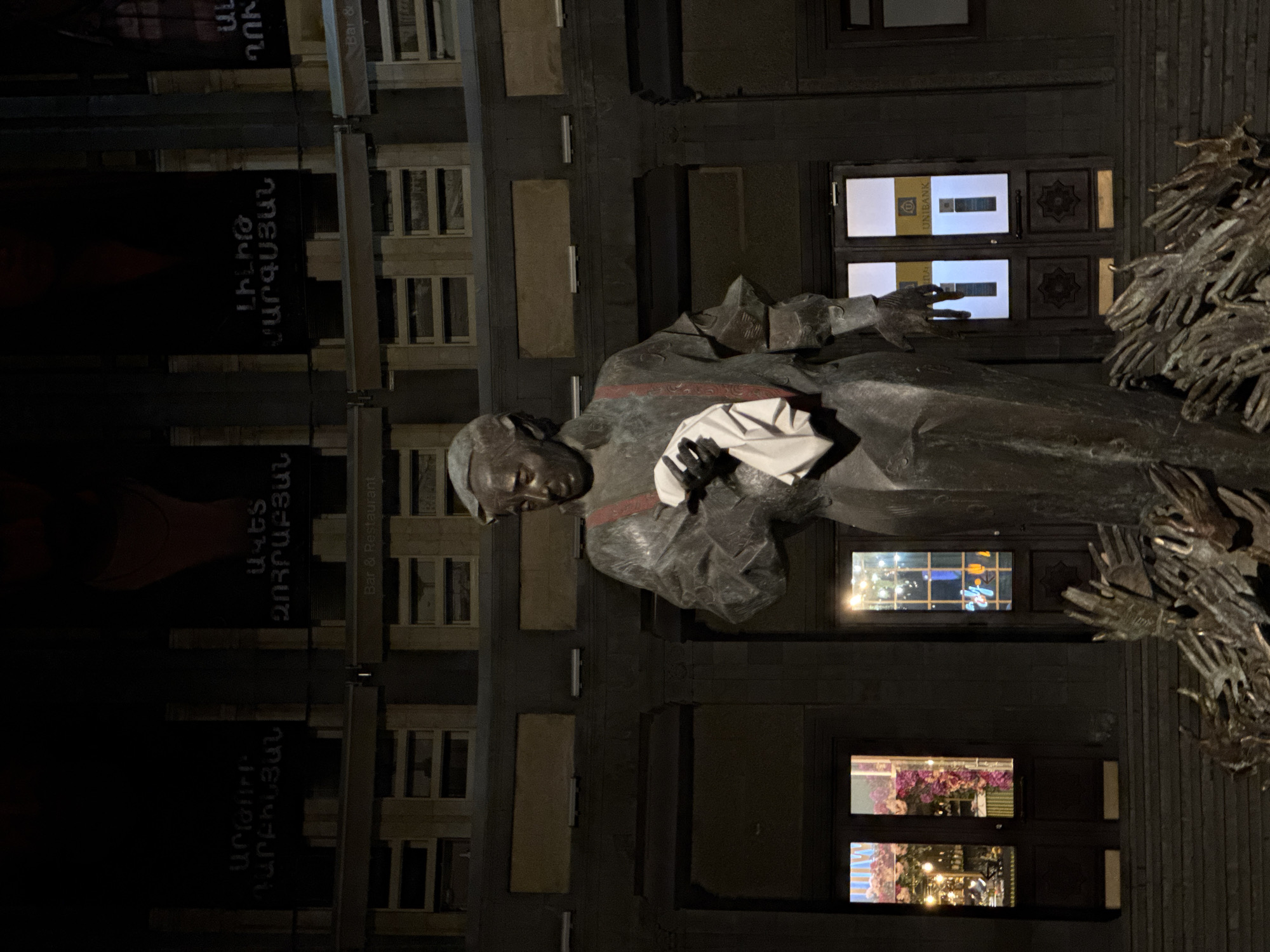 A bronze statue of a seated man, presumably a writer or intellectual, dominates the frame, set against the backdrop of a multi-story building at night. The statue is likely the Yegishe Charents monument, positioned in front of the Yerevan State University of Architecture and Construction in Yerevan, Armenia. The man in the statue is dressed in a cloak or gown and is holding a white object, potentially a book or paper. Behind the statue, on the right side, is a structure made of many outstretched metal hands. The building facade is visible with numerous windows, some of which are illuminated, reflecting activity inside. Dark banners or signs with text hang from the building, partially obscuring the view. The overall lighting is dim, emphasizing the statue as the central subject.
