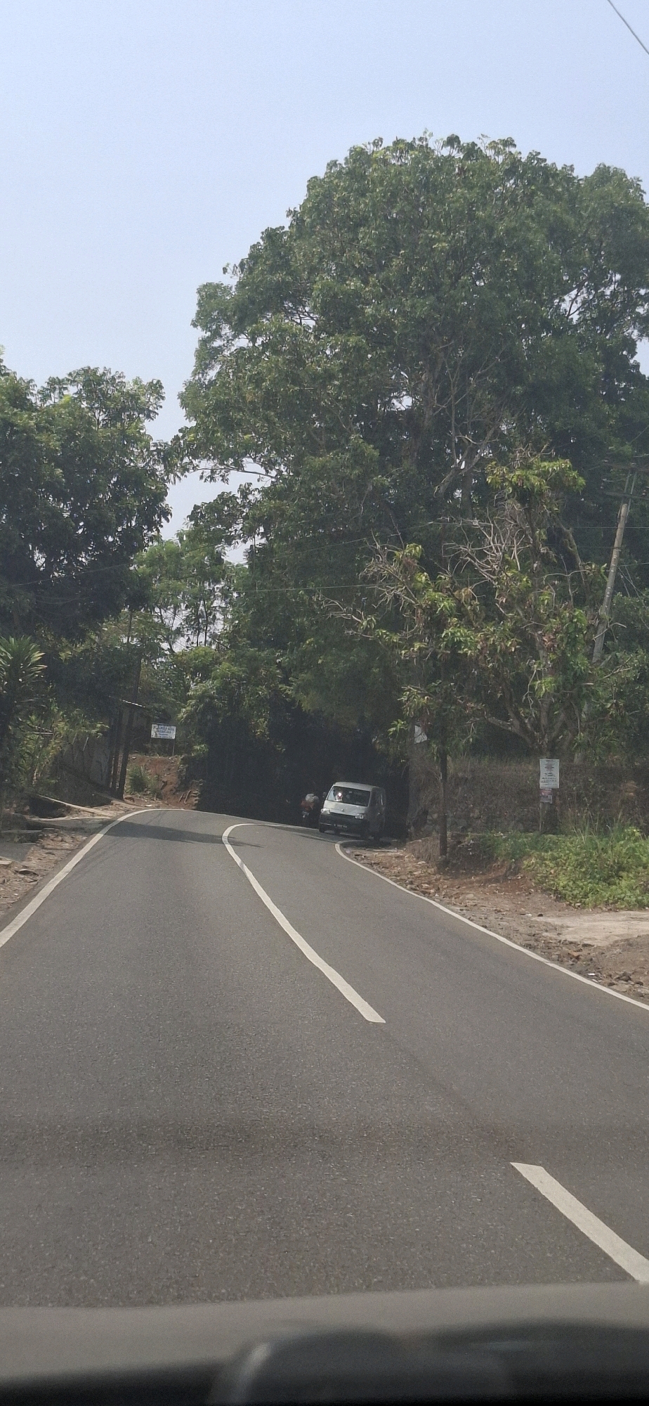 The image captures a winding asphalt road passing through a lush, green environment. Tall trees with dense foliage line both sides of the road, creating a shaded canopy in some sections. The sky above is a pale blue, with a hint of haze. A dark-colored van is parked on the right side of the road, partially obscured by vegetation. In the distance, on the left side of the road, a small sign is visible, mounted on a pole. The road surface is well-maintained, with a clear white line marking the center. The overall impression is a tranquil, rural scene, likely in Susukan, Indonesia, given the context.