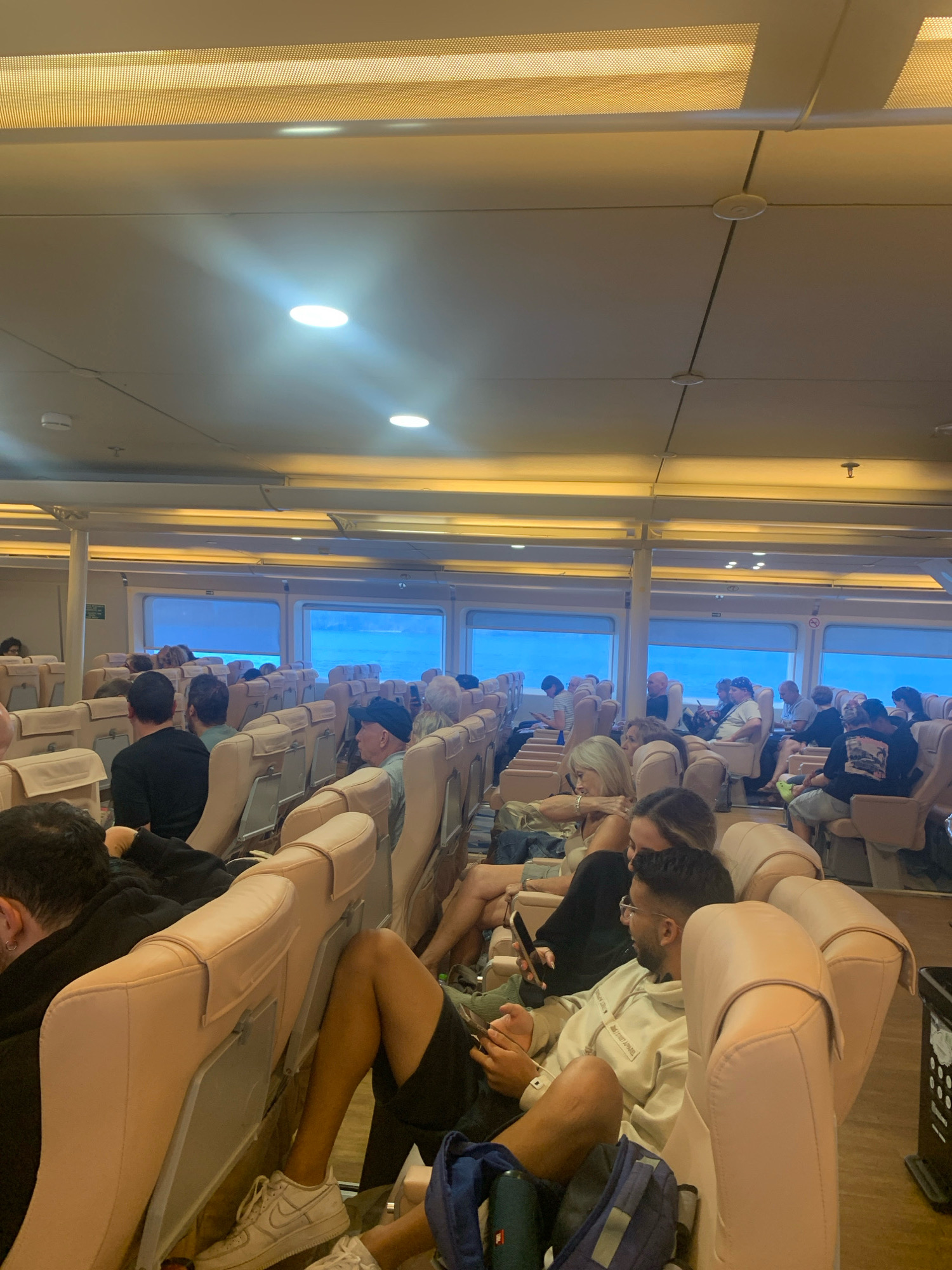 The image shows the interior of a passenger ferry, likely en route from Glossa, Greece, given the context. The cabin is filled with rows of beige upholstered seats, many of which are occupied by passengers. The lighting appears to be artificial, with fluorescent strips visible along the ceiling. Large windows line the sides of the cabin, offering a view of the blue sea and sky. Passengers are dressed casually, and some are engaged with their phones or looking out the windows. The atmosphere appears relaxed, consistent with a ferry journey.