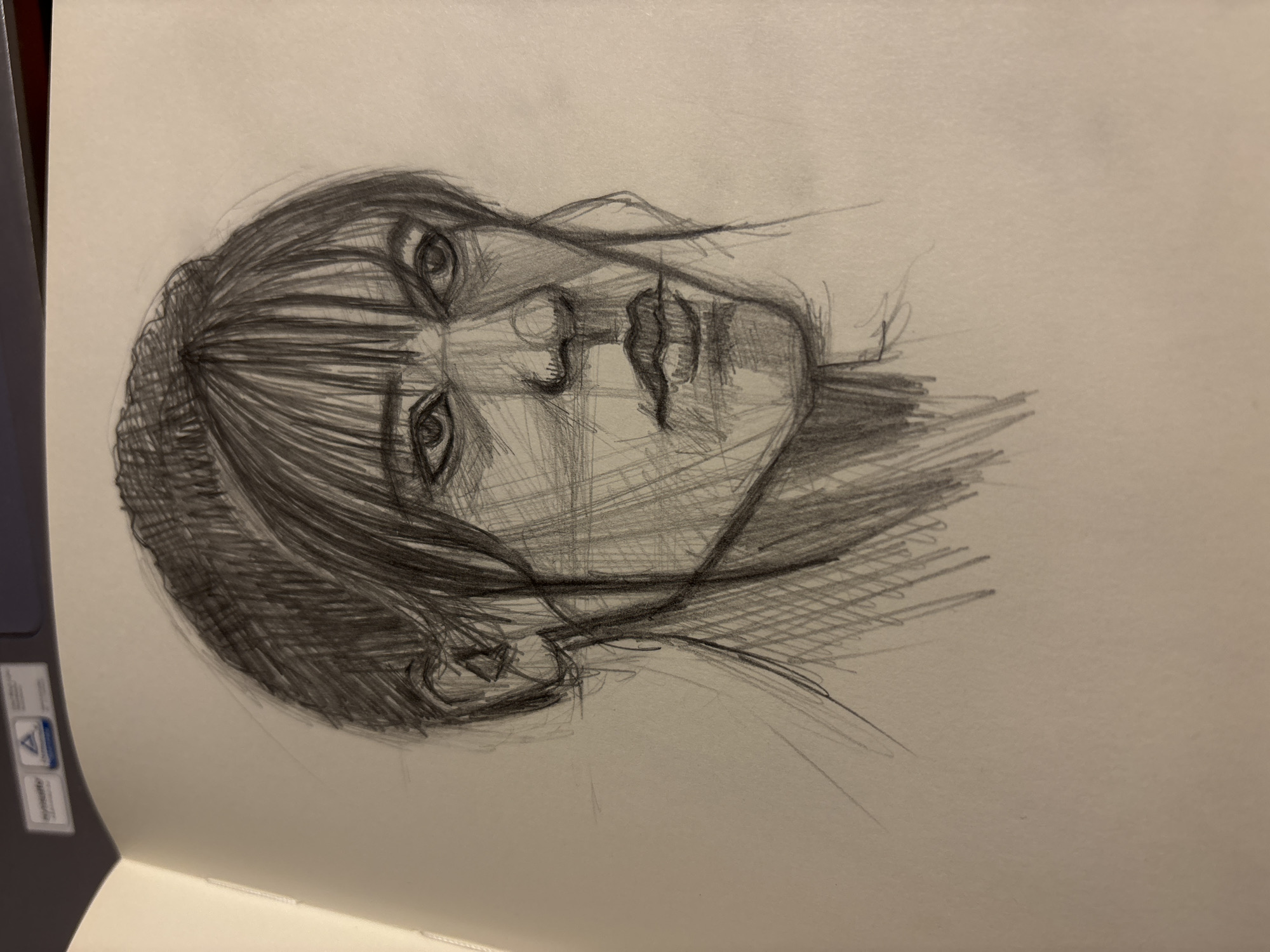 The image shows a pencil sketch of a person's face. The subject has bangs and is angled towards the viewer's right. Details include shading on the hair, face, and neck. The drawing is on a piece of paper within a notebook. Part of a laptop is visible in the lower left corner. There is no indication of Izhevsk, Russia.