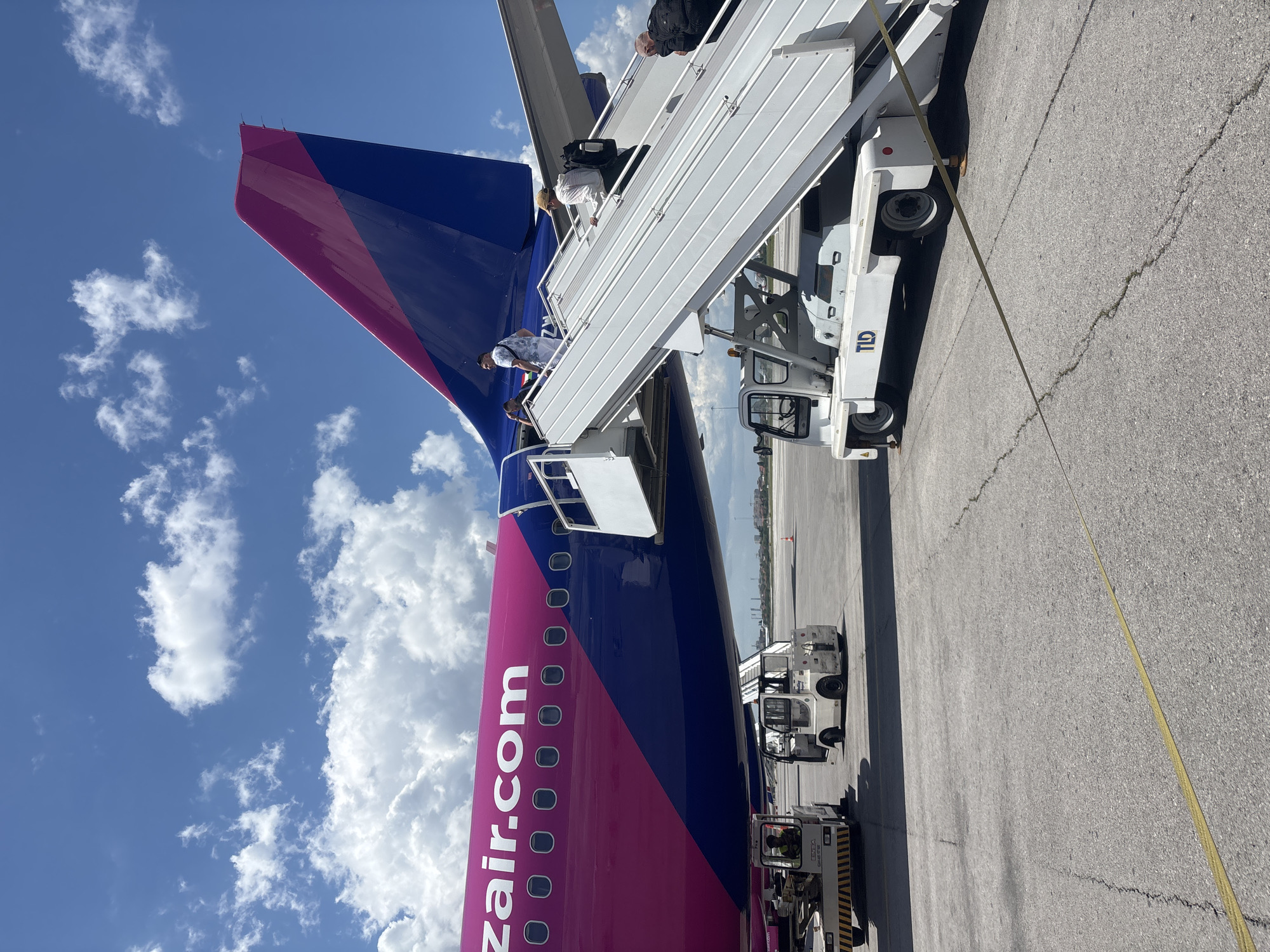 An airplane with the Wizz Air logo and "wizzair.com" written on its fuselag is parked on an airport tarmac. Passengers are seen boarding the aircraft via a portable airstair. Two baggage tugs are positioned near the plane, one on the left and another on the right. The scene takes place on a sunny day with a blue sky and scattered white clouds, characteristic of an outdoor airport environment in Sofia, Bulgaria.