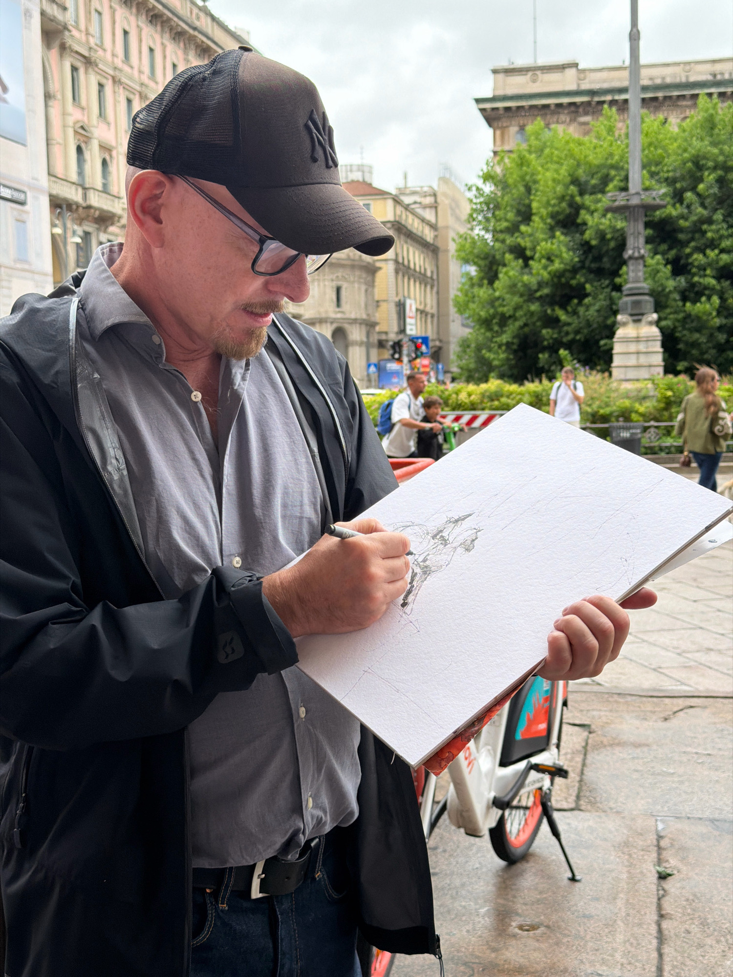 In Milan, Italy, a man wearing a black New York Yankees baseball cap and glasses is captured in a medium shot as he draws on a white sketchpad. He is dressed in a gray collared shirt under a black rain jacket and blue jeans, cinched with a black leather belt. The man's left hand holds the sketchpad steady, while his right hand uses a pen to sketch. The sketchpad, open on a white clipboard, reveals a detailed drawing of a horse in progress. In the background, blurred figures walk along a paved city street, with buildings and lush green trees suggesting an urban environment. A red and white bicycle with an orange wheel is partially visible on the right side of the frame. The sky is overcast, indicating a cloudy day.