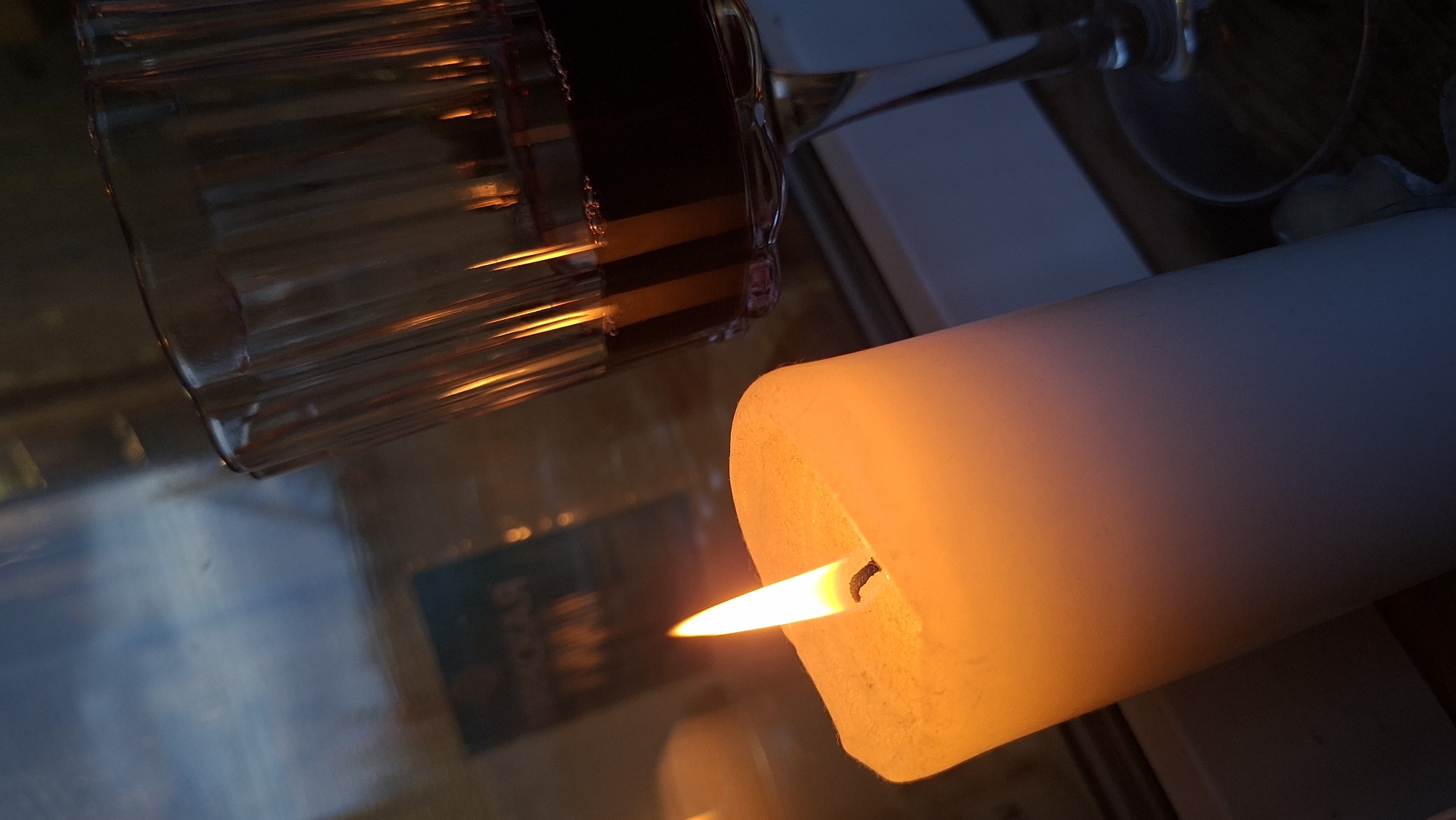 The image shows a lit candle and a glass of red wine. The candle is on the left side of the frame, with its flame brightly illuminating the scene. To the right of the candle is a glass of red wine, partially filled. The background appears to be a window with reflections of the outdoors and some text that is blurred and illegible. The lighting suggests it is evening or nighttime. The scene is indoors, likely in a home or a dimly lit establishment. There is no information to indicate the location in Забалоцкі сельскі Савет, Belarus.