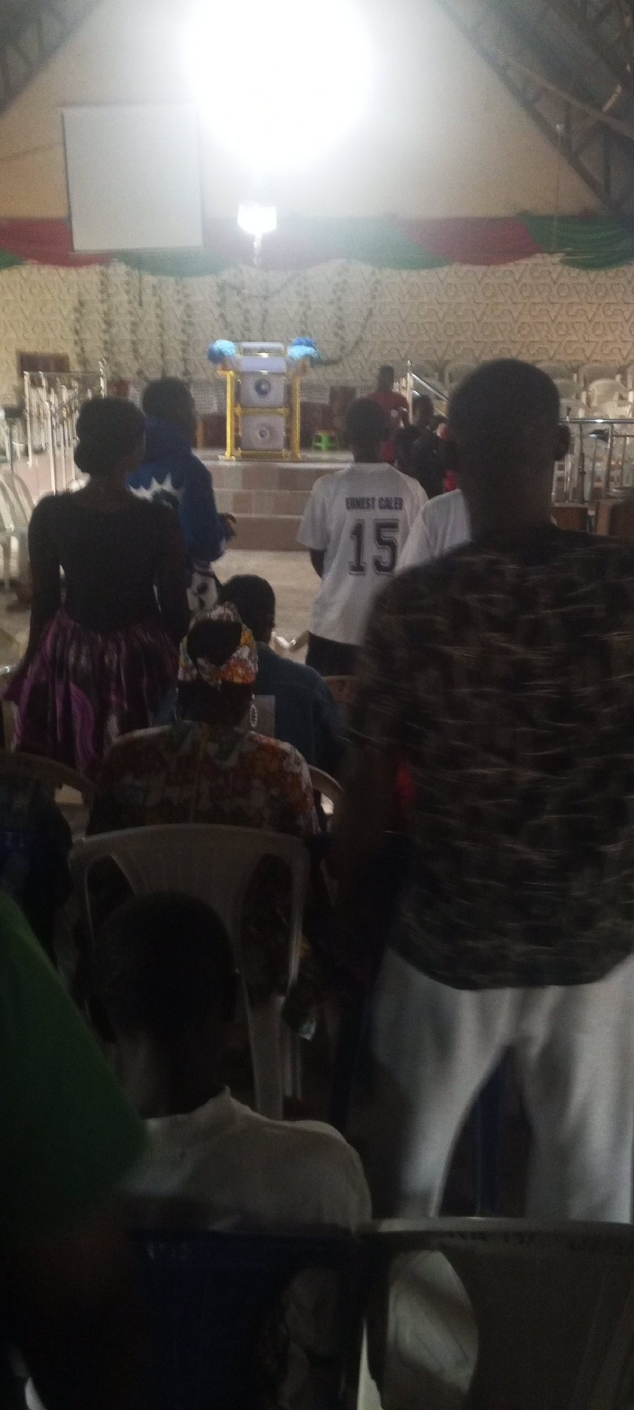 The image captures an indoor scene, possibly a church or a community hall in Bahit, Nigeria. The lighting suggests the presence of bright overhead lights. In the foreground, the backs of several individuals are visible, with one person in a white t-shirt with the number "15" and the name "ERNEST GALER" printed on it. To the left, a person in a black top and a patterned skirt is seen from behind. In the middle ground, a podium or stage area is visible, decorated with blue and yellow accents and featuring what appear to be speakers. The background shows more people, chairs, and a wall decorated with a patterned fabric and draped fabric in red and green.