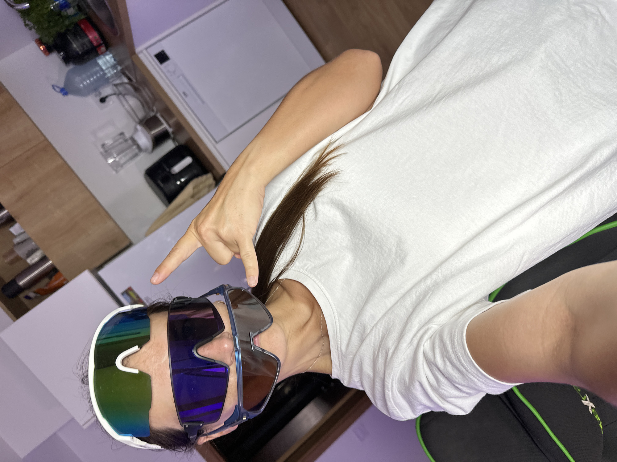 A medium shot shows a person wearing multiple pairs of sunglasses layered on their face, covering most of their eyes and nose. The person is wearing a white t-shirt and has long brown hair. Their right hand is raised, with the index finger pointing upwards and the thumb and pinky extended in a gesture. The background appears to be a kitchen, with cabinets, a blender, a water bottle, and a dishwasher visible. The lighting is dim with a purple hue in the background. There is no visible indication of the location in Donji Pobori, Montenegro.