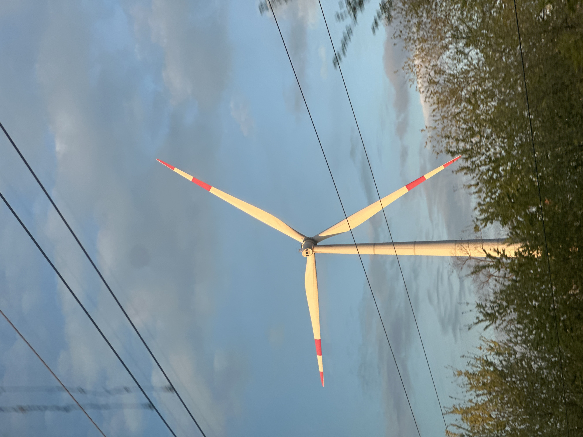 The image shows a wind turbine with three blades, each tipped with red and white stripes. The turbine is positioned against a sky with scattered clouds. Power lines run across the lower-left portion of the frame. The right side of the image features the foliage of trees. The specific location in Zimovniki, Russia, cannot be determined from the image.