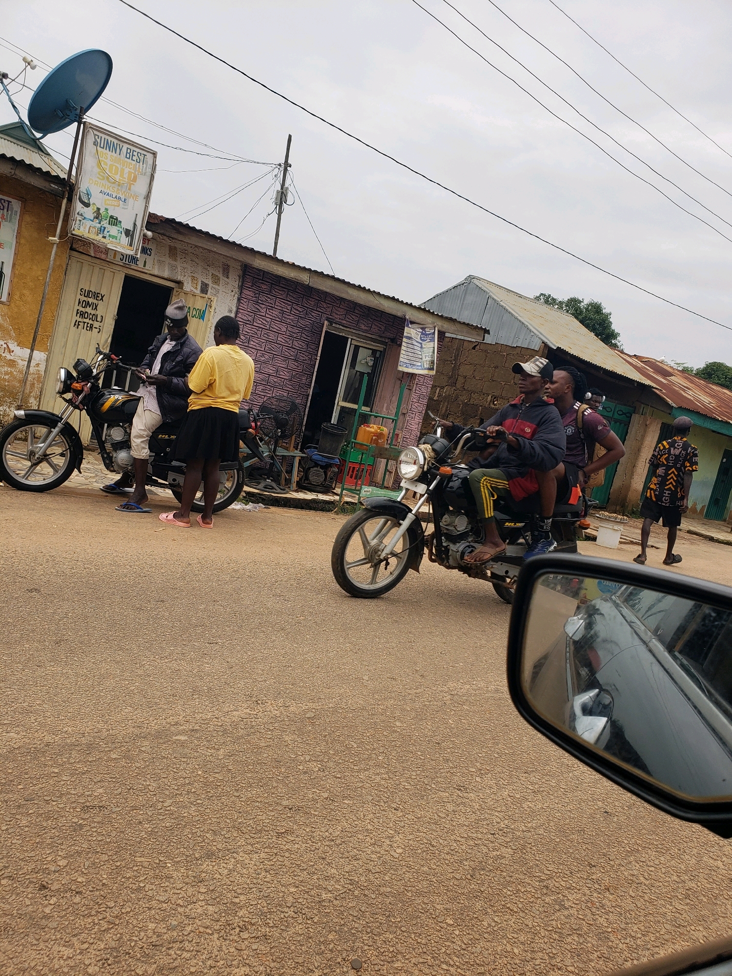 In Narayi, Nigeria, on an overcast day, three individuals ride on two motorcycles, one with two passengers and the other with one, while a fourth person stands on the side of the road. The motorcycles are on a paved road in front of a row of shops. The shop on the left has a blue satellite dish on its roof and a sign that reads "SUNNY BEST." The middle shop has a display of bottled drinks and wine. The shop on the right has a corrugated metal roof. The road is lined with power lines and poles. The overall scene depicts a typical day in a Nigerian city, with people going about their business.