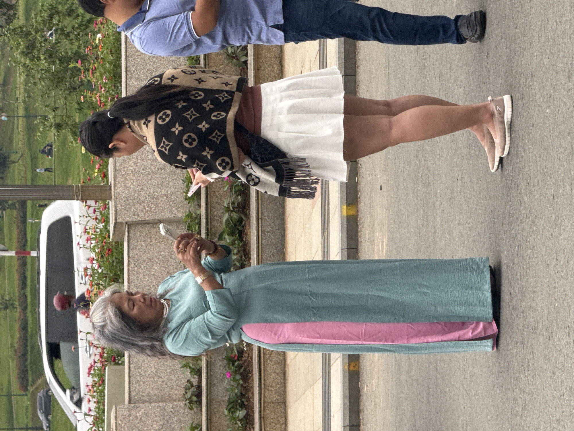 In Phường Xuân Hương, Đà Lạt, Vietnam, three individuals are visible on a paved street in front of a textured stone wall with flowerbeds. A woman with gray hair, wearing a teal dress with a pink insert and a pearl necklace, stands facing left, holding a smartphone. To her right, a woman with her dark hair in a ponytail is seen from the back, wearing a white pleated skirt and a patterned scarf with a Louis Vuitton-like design over a brown top. She is also holding a smartphone. To the right of these two women, a man in a blue polo shirt and dark jeans is partially visible, facing away from the camera. The background includes a white vehicle, greenery, and other people in the distance.