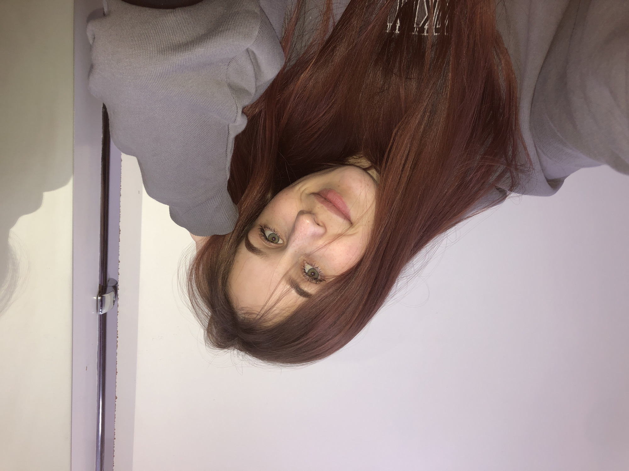 The image displays a close-up portrait of a female subject in an indoor setting. The subject has long, reddish-brown hair, light-colored eyes, and a visible scattering of freckles across her nose and cheeks. She exhibits a slight, closed-mouth smile and is looking directly at the viewer. She is wearing a light grey hooded sweatshirt. Her left arm is bent, with her hand positioned near her temple, partially supporting her head. The background consists of a plain, uniformly lit light-colored wall. To the right of the subject, a section of a white door frame is visible, including a chrome-plated door latch mechanism. A faint shadow is present on the far right section of the wall. The scene is located in the city of Фоки, Russia.