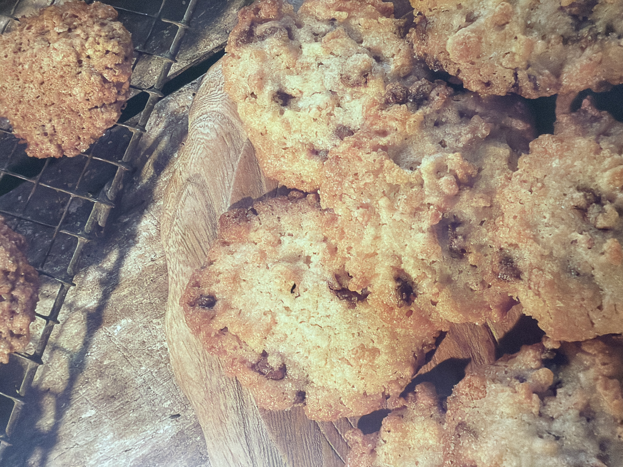 The image shows a close-up, overhead view of several golden-brown, textured cookies, some on a rustic wooden surface and others on a metal cooling rack. The cookies appear to be homemade, with visible chunks of what might be chocolate or nuts embedded within their crumbly texture. The lighting creates shadows that highlight the uneven surfaces of the cookies and the grain of the wood. One cookie is prominently displayed on the cooling rack in the upper right portion of the image, while several others are clustered on the wooden surface in the foreground. The overall impression is one of warmth and deliciousness.