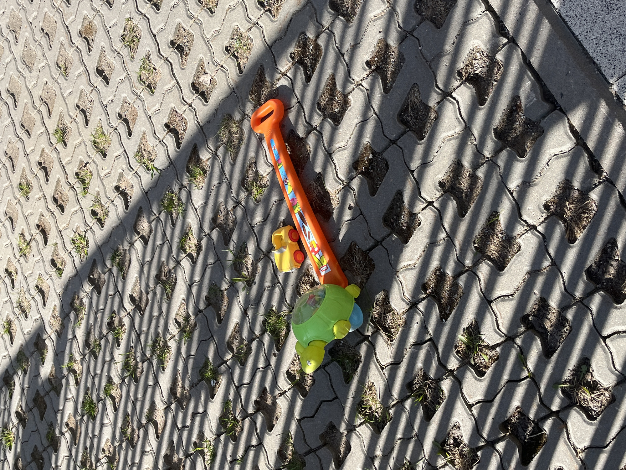The image displays a ground-level view of an outdoor paved area, featuring permeable concrete pavers. These pavers have a patterned, interlocking design with diamond-shaped openings, many of which contain small tufts of green grass or dry soil.
Two children's toys are prominently placed on the pavement. The main subject is an orange push toy, resembling a child's lawnmower or similar, with a large handle at the top and colorful, cartoon-like decals along its body. Adjacent to it is a smaller, green turtle-shaped toy with yellow accents, including wheels, and a clear dome on its back. A small, yellow toy car is partially visible near the base of the orange push toy.
Distinct, long, parallel shadows are cast diagonally across the entire paved surface, suggesting bright sunlight and indicating either morning or late afternoon. These shadows likely originate from an unseen fence, railing, or similar structure. The weather appears sunny.
The scene depicts an ordinary outdoor setting, possibly a pathway, a residential area, or a children's play space, where toys have been left unattended. No people are visible. The colorful plastic toys stand out against the grey concrete and natural elements. While there are decals on the orange toy, any text present is not legible due to the resolution and angle.