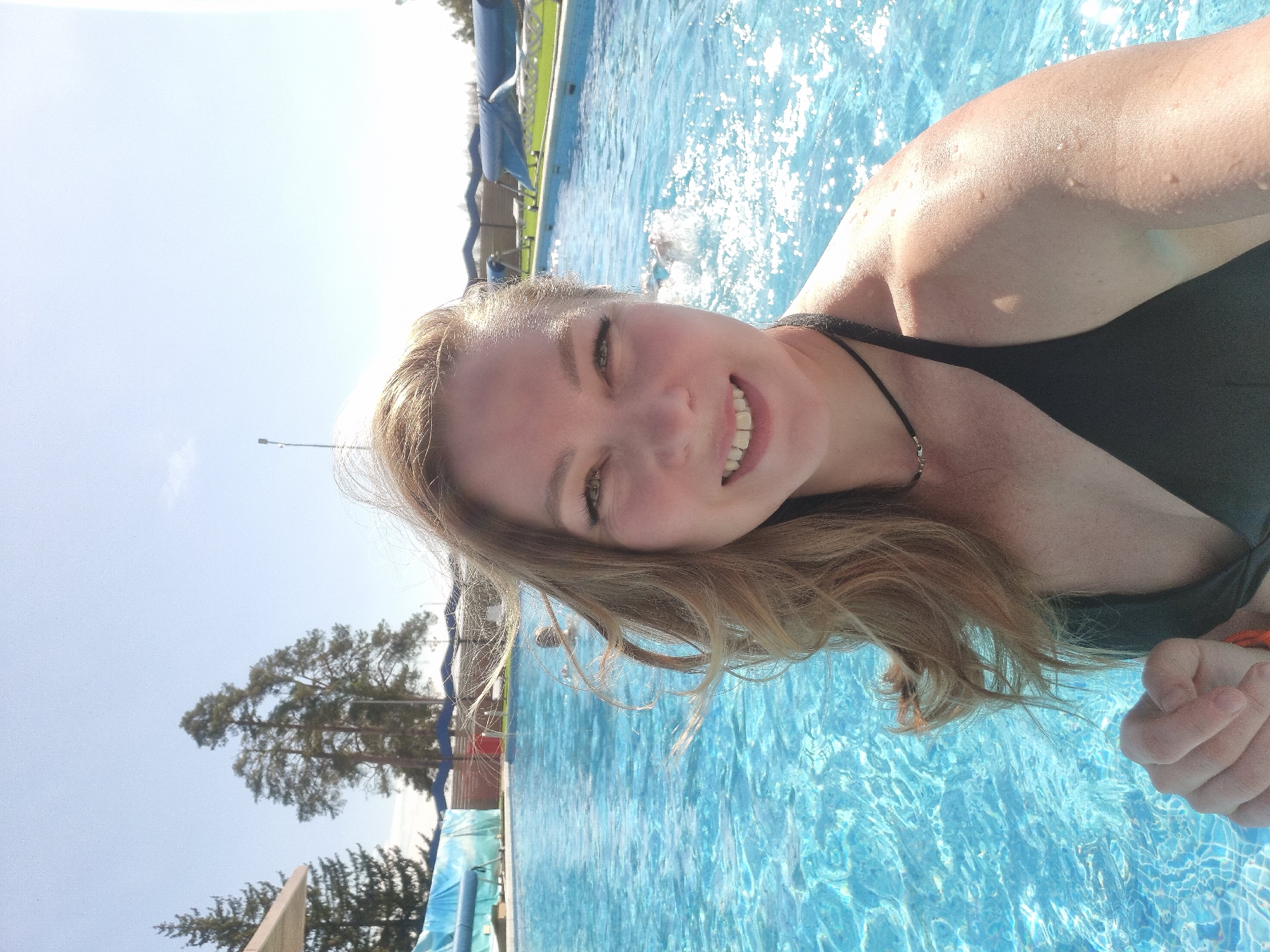 A selfie-style, rotated, medium shot depicts a smiling woman in a swimming pool. Her face and upper right shoulder are visible, wet with water droplets. She wears a black swimsuit with a thin necklace. Her hair is blonde. The pool has clear blue water with sunlight reflecting on the surface. In the background are a tree and a building. The location is Votkinsk, Russia.