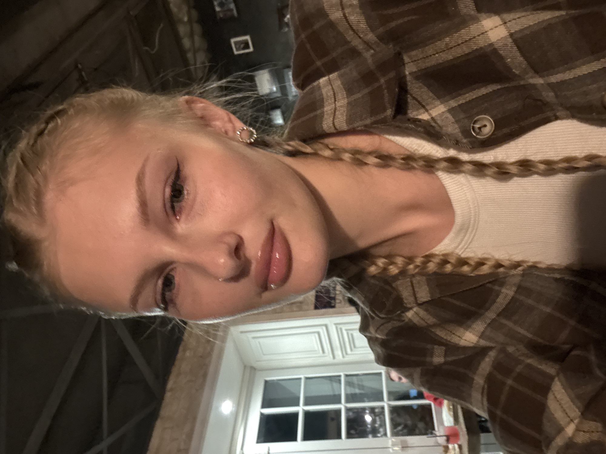 A young woman with blonde hair styled in two braids and a light brown plaid shirt is looking at the camera. She is wearing a white ribbed tank top underneath her shirt, and a small circular earring in her right ear. The background suggests an indoor setting, possibly a cafe or restaurant, with a window and a ceiling fan visible. The lighting is dim. It is not possible to determine the location of the city of Perm, Russia from the image alone.