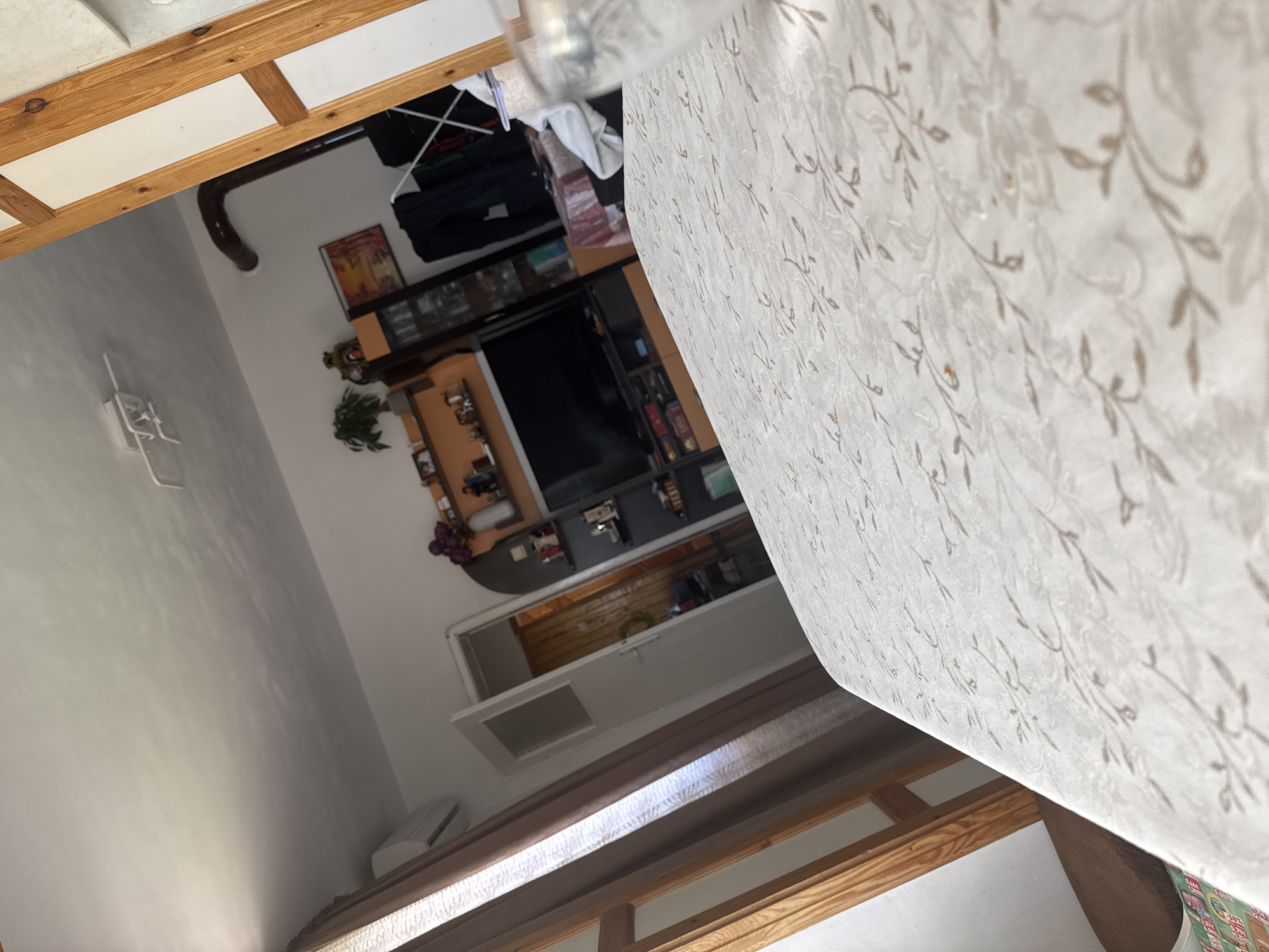 The image shows an interior scene, likely a home, with a TV and entertainment unit against a wall. A doorway is visible, leading to another room. A patterned white and beige tablecloth dominates the foreground. The location is Topoli, Bulgaria, although specific location details within Topoli are absent. There are no people visible in the image.