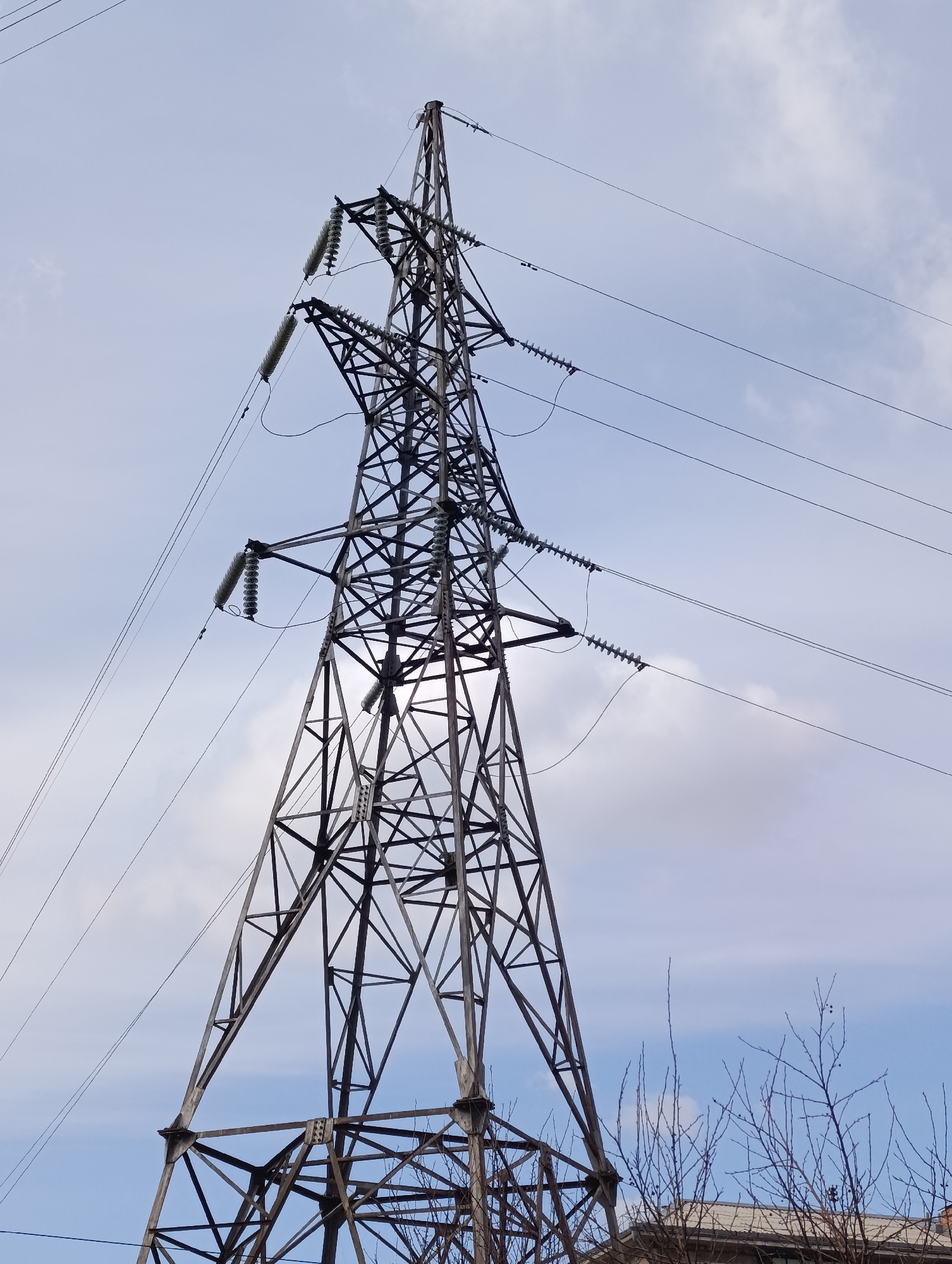 The image shows a high-voltage transmission tower against a cloudy sky. The tower is made of a lattice of metal beams and has multiple insulators supporting electrical wires. The base of the tower is partially obscured by foliage and a building. The image was taken in Krasnoyarsk, Russia.