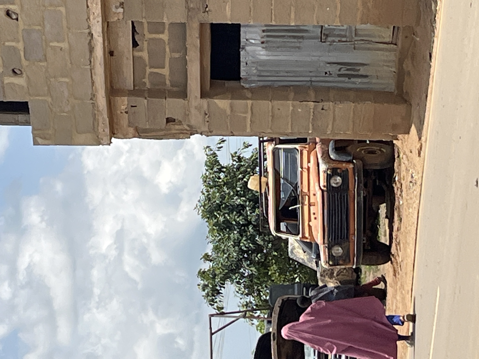 In Rafin Par, Nigeria, an orange off-road vehicle is parked on a dirt road next to a partially constructed building made of cinder blocks. A person in a pink headscarf and blue pants walks away from the camera on the left side of the road, passing an open vehicle. Behind the orange vehicle, there is a dense green tree and a metal frame structure. The sky above is filled with white and gray clouds.