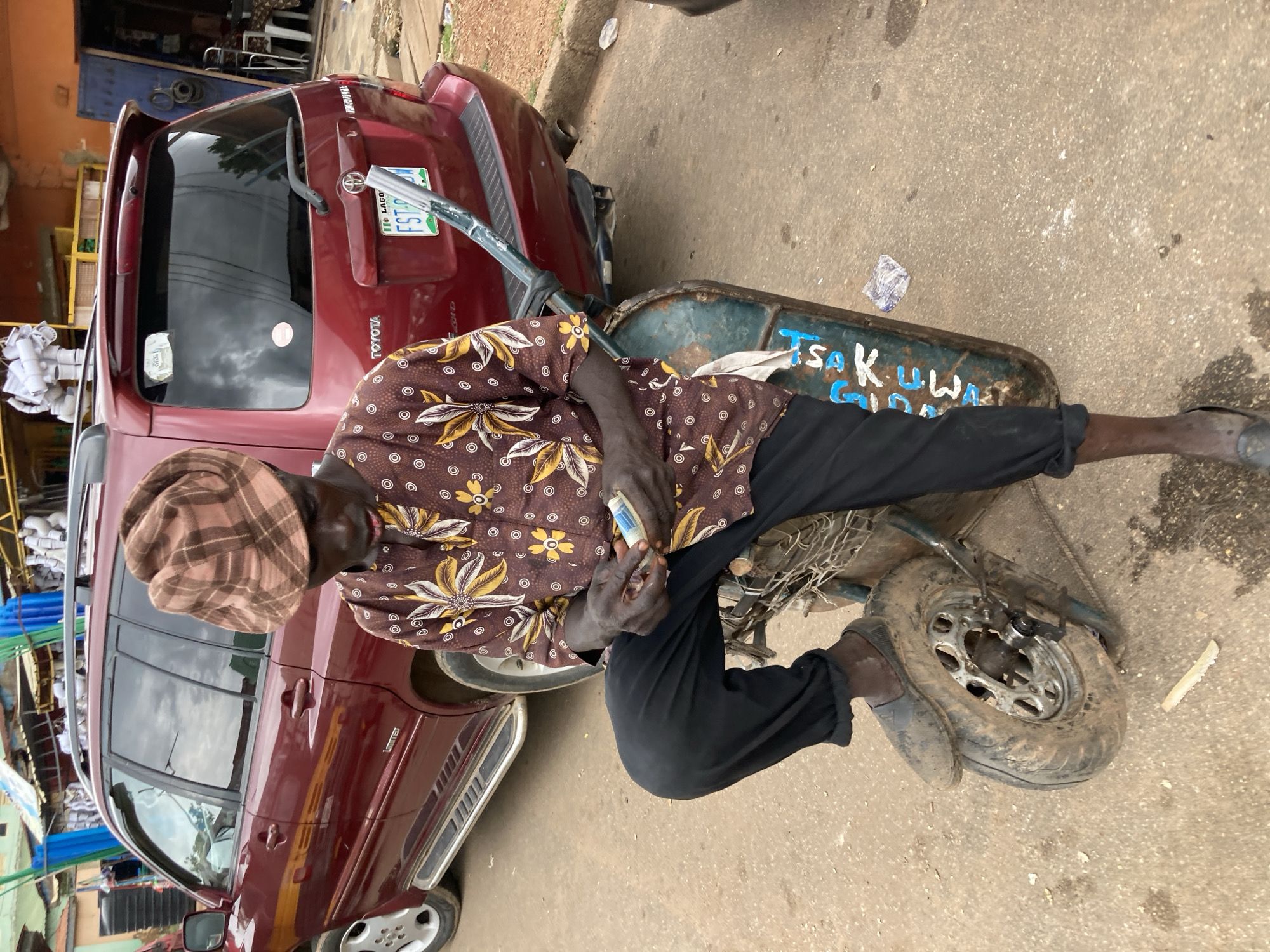 The image shows a person sitting on a handcart next to a maroon Toyota SUV. The person is wearing a brown, floral-patterned shirt and black trousers, with a checkered headscarf. Their hands are visible, holding what appears to be a packet of cigarettes. The handcart is green and has some blue lettering on its side. The SUV is parked on a paved surface, possibly a street or market area, with a curb and some debris visible on the ground. In the background, there are hints of buildings and other vehicles, suggesting an urban or semi-urban environment. The lighting suggests it is daytime, and the scene appears to be set in Jos, Nigeria. There is no clear indication of the weather or any strong emotions conveyed by the subject. Visible text includes "TOYOTA" on the back of the SUV and "FST 290 EQ" on a license plate. The blue lettering on the handcart is partially legible, reading "TSAKUWA".